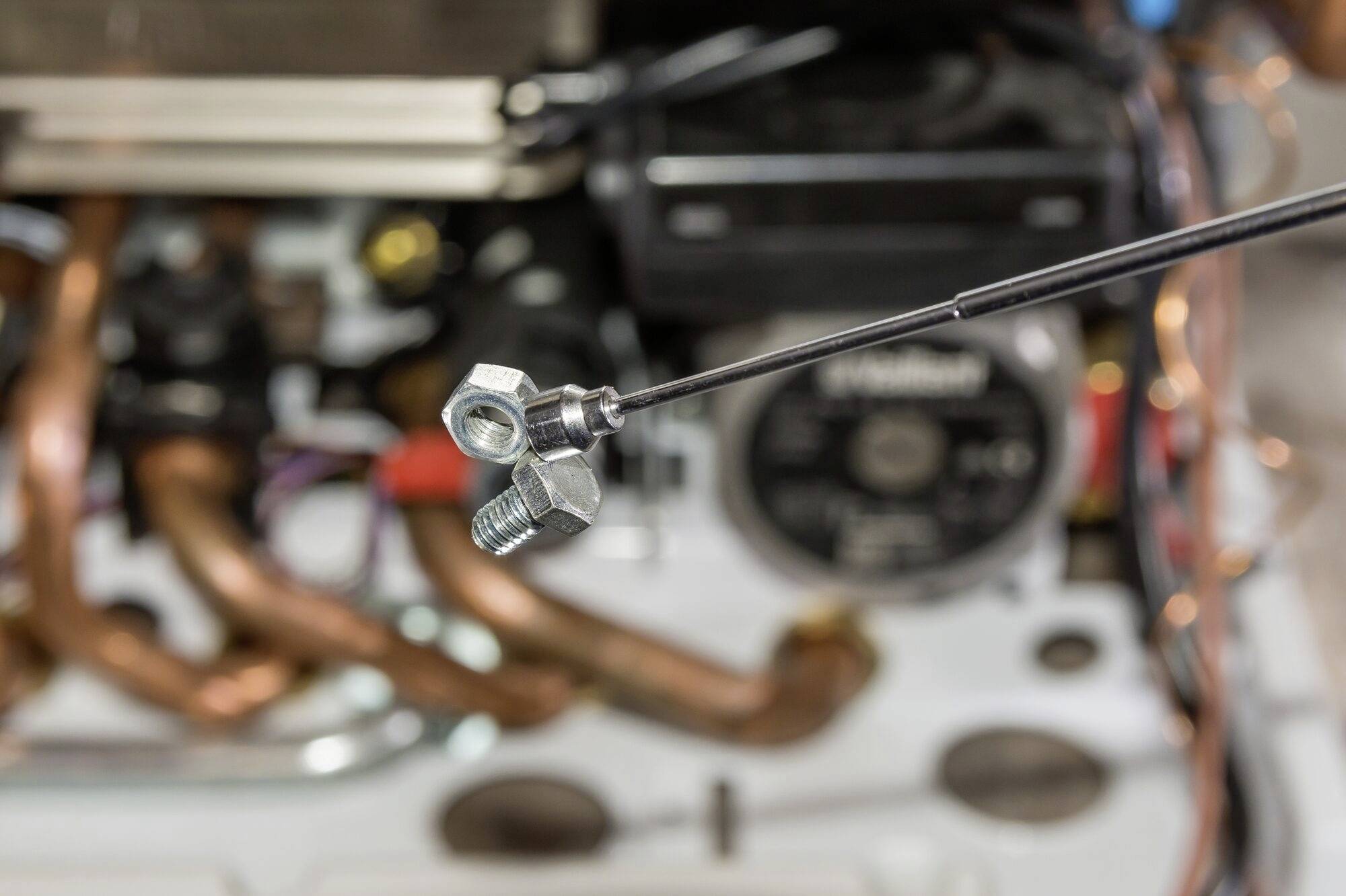 Detailný záber magnetického uchopovača držiaceho matice a skrutky pred rozmazaným pozadím medených rúrok a strojových súčiastok.