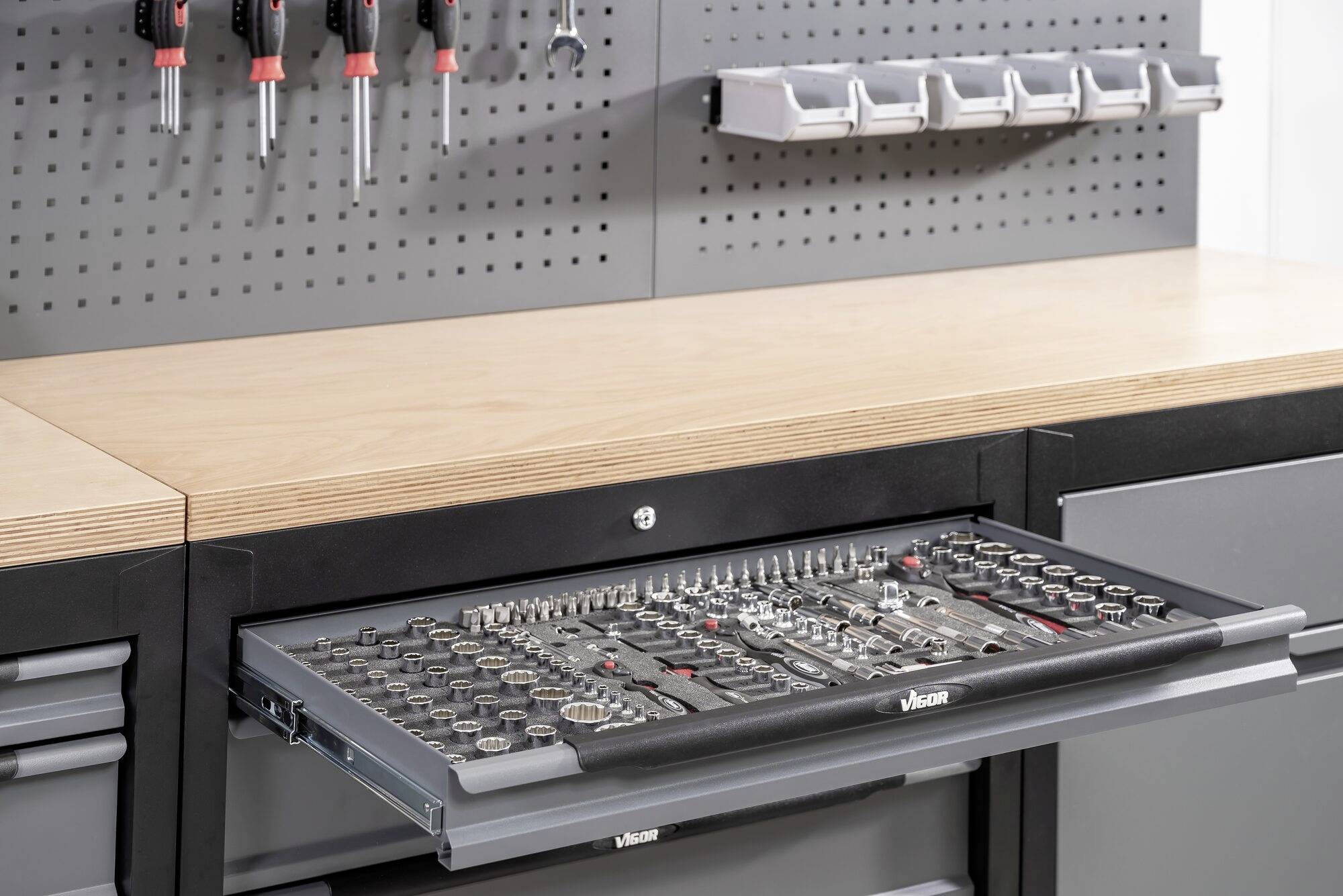 Zásuvka na náradie v pracovnom stole, usporiadaná so sadami nástrčných kľúčov a nástrojmi; nad ou skrutkovače zavesené na dierovanej stene.