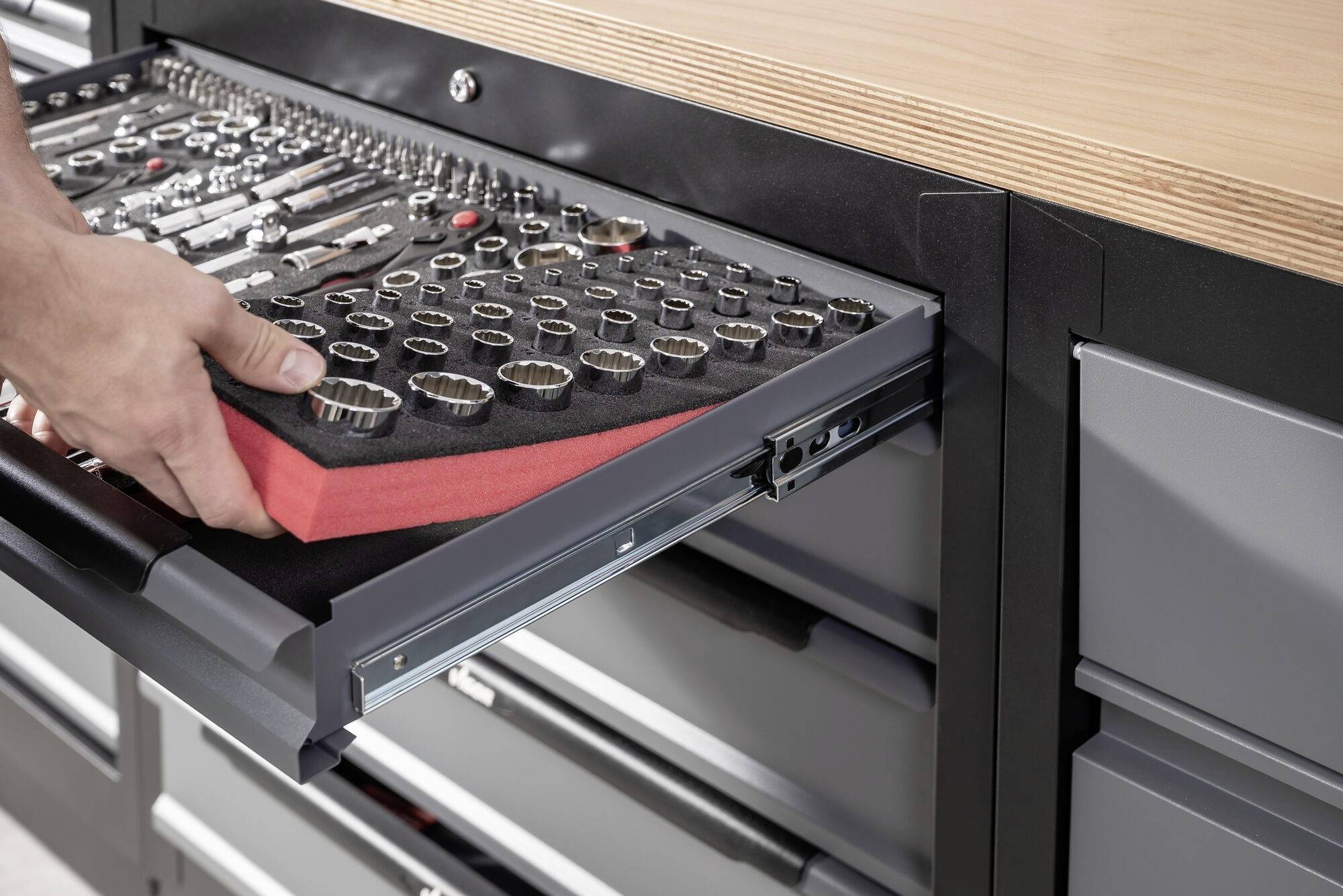 'Vyťahovateľný zásuvkový organizér s utriedenými nástrčkovými kľúčmi je vyťahovaný jednou rukou. V pozadí je pracovný stôl s drevenou doskou.'