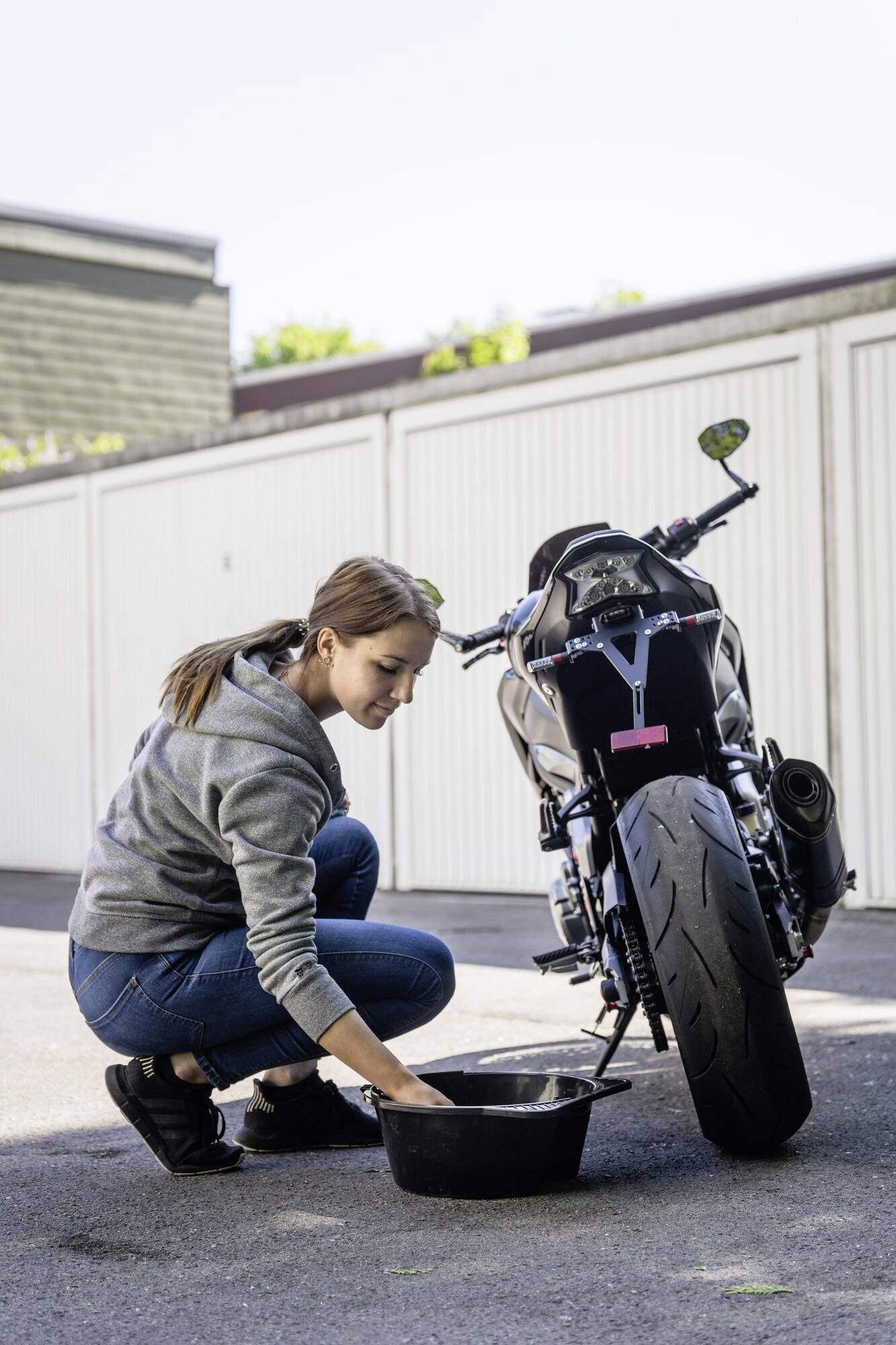 Žena v sivej mikine sedí kľačmo vedľa motocykla na dvore. Pracuje s čiernou nádobou, pravdepodobne na údržbu motocykla.