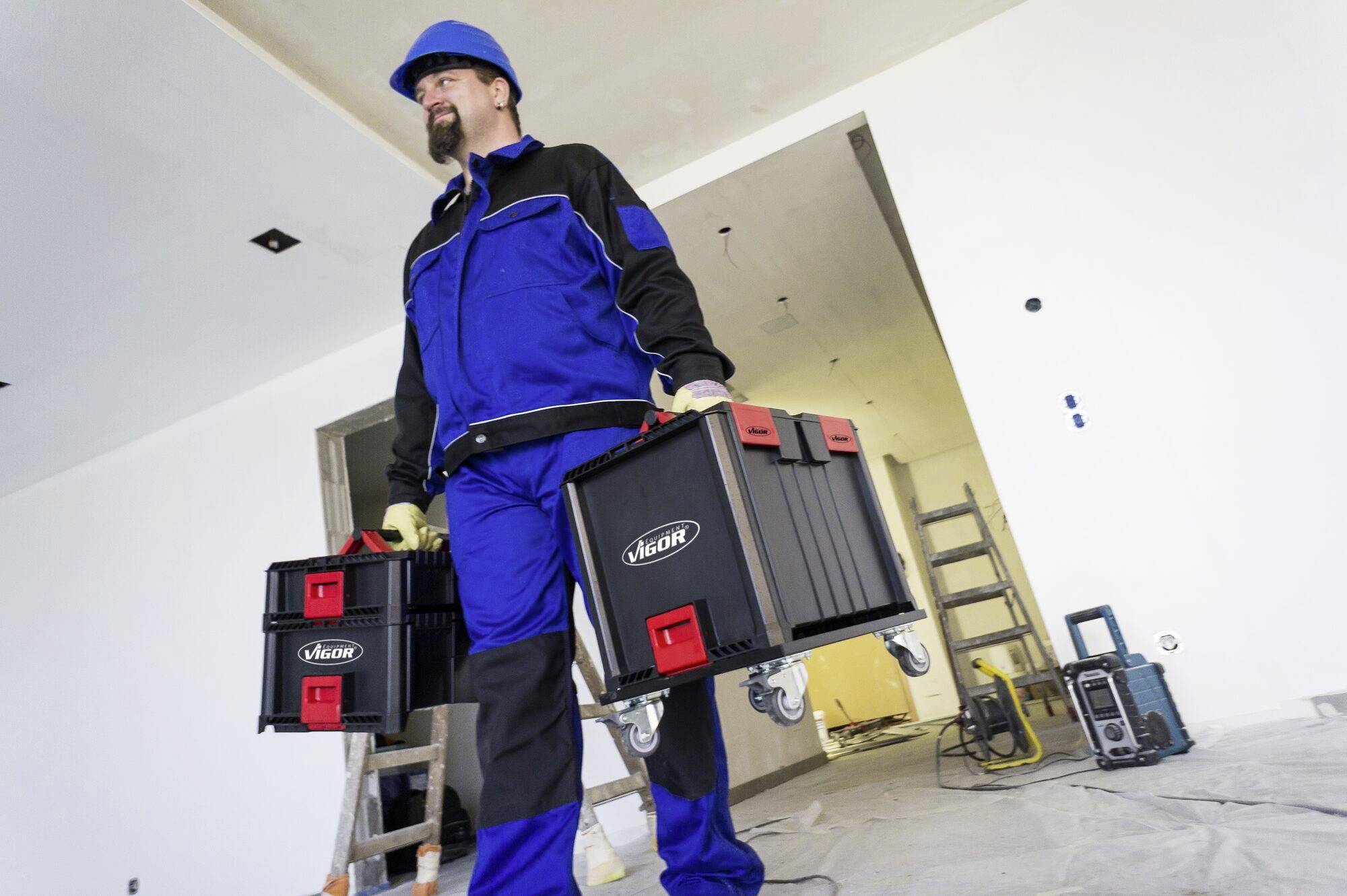 Robotník v modrom ochrannom oblečení a s prilbou nesie dve debny s náradím v nedokončenej miestnosti obklopenej stavebným zariadením.