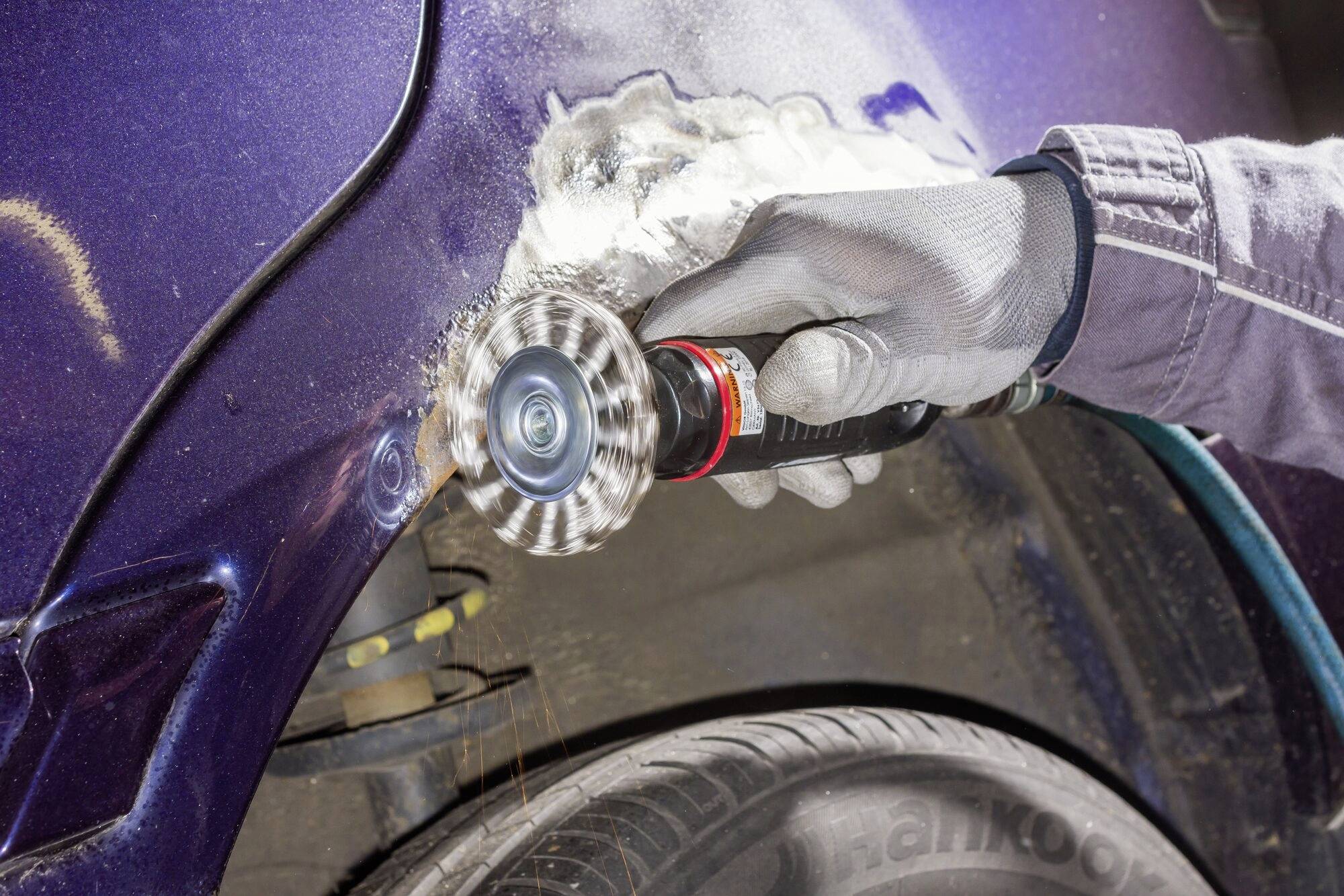 Mechanik brúsi hrdzu z modrého autokríidla uhlovou brúskou. Od brúsenia odlietavajú iskry, pracovník má oblečený ochranný pracovný odev.