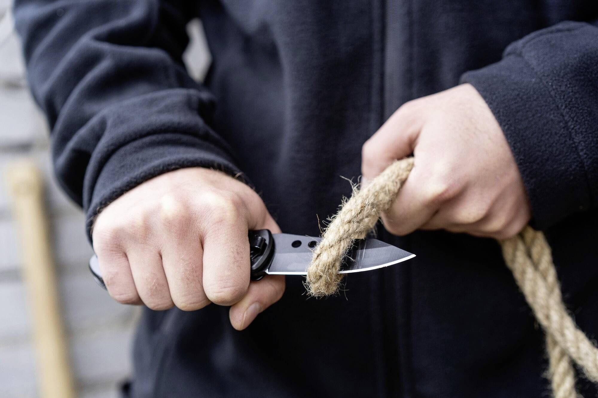 Muž v čiernej bunde strihá lano nožom. Detailný záber ukazuje ruky a samotné strihanie.