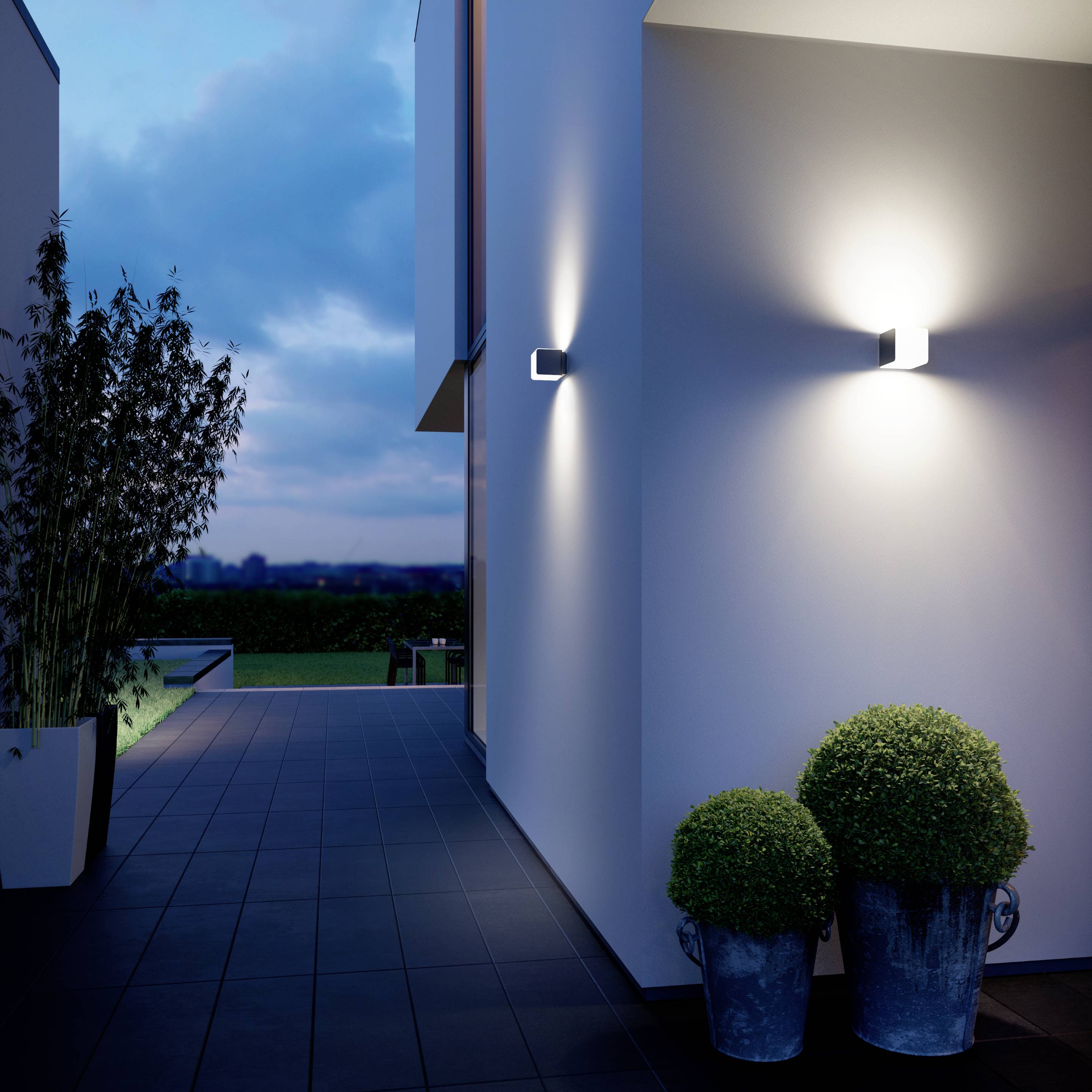 Moderný vonkajší priestor za súmraku, osvetlený stenový svietidlami. Chodník vedie medzi bielymi budovami, ozdobenými rastlinami.