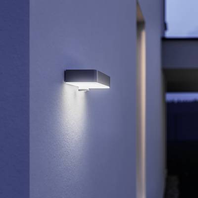 Moderná vonkajšia lampa na stene domu svieti v súmraku. Poskytuje svetlo na fasádu a chodník.