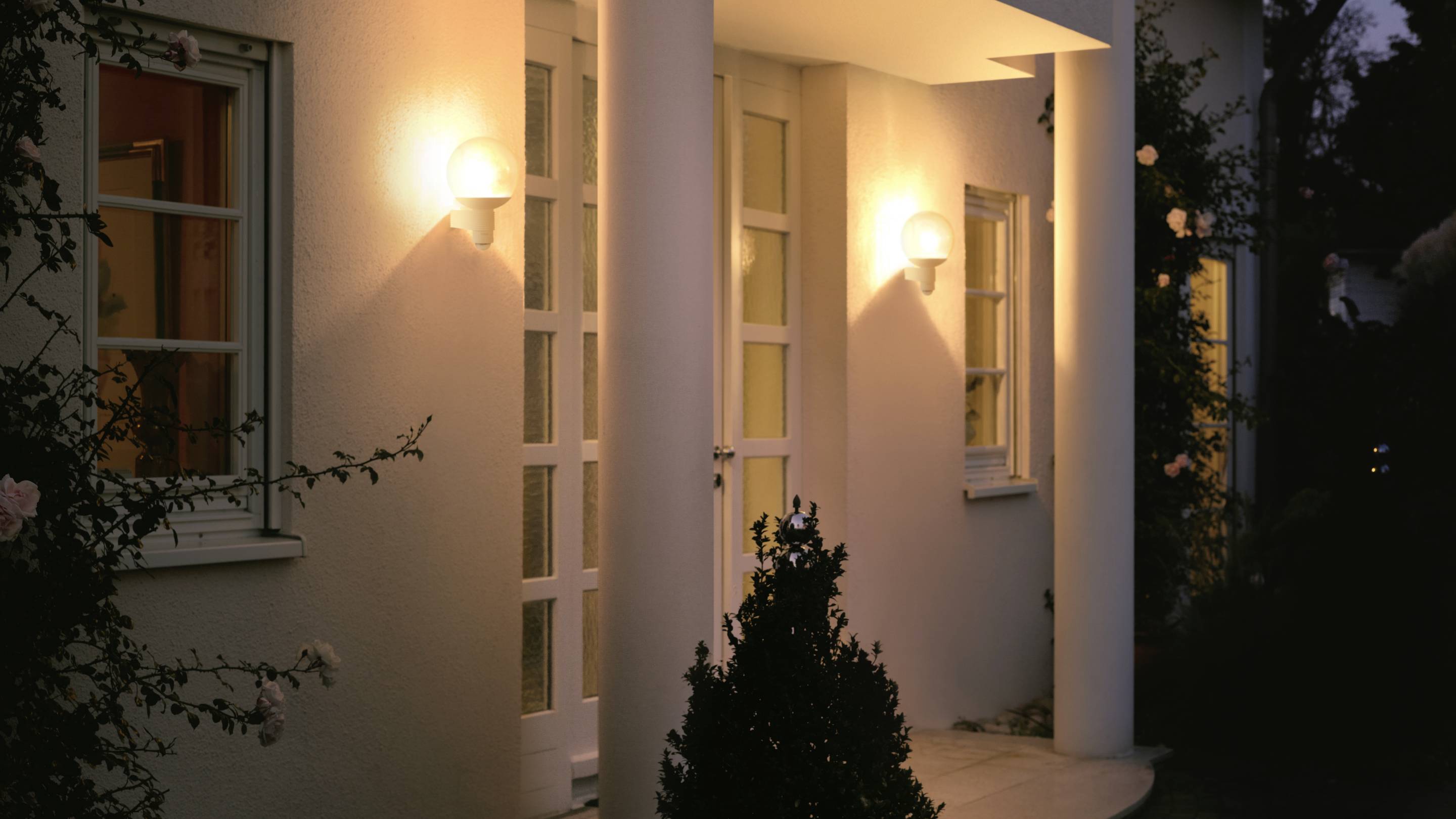 Vstup rodinného domu v noci, osvetlený dvoma nástenými lampami, s bielymi stĺpmi a kríkom v popredí.