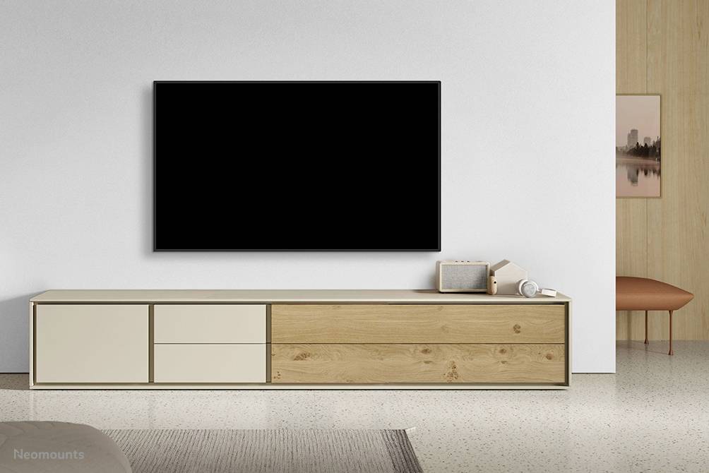 Plochá televízia na bielej stene, pod ňou minimalistická drevená komoda v svetlo béžovej farbe s tromi zásuvkami a dvomi dvierkami.