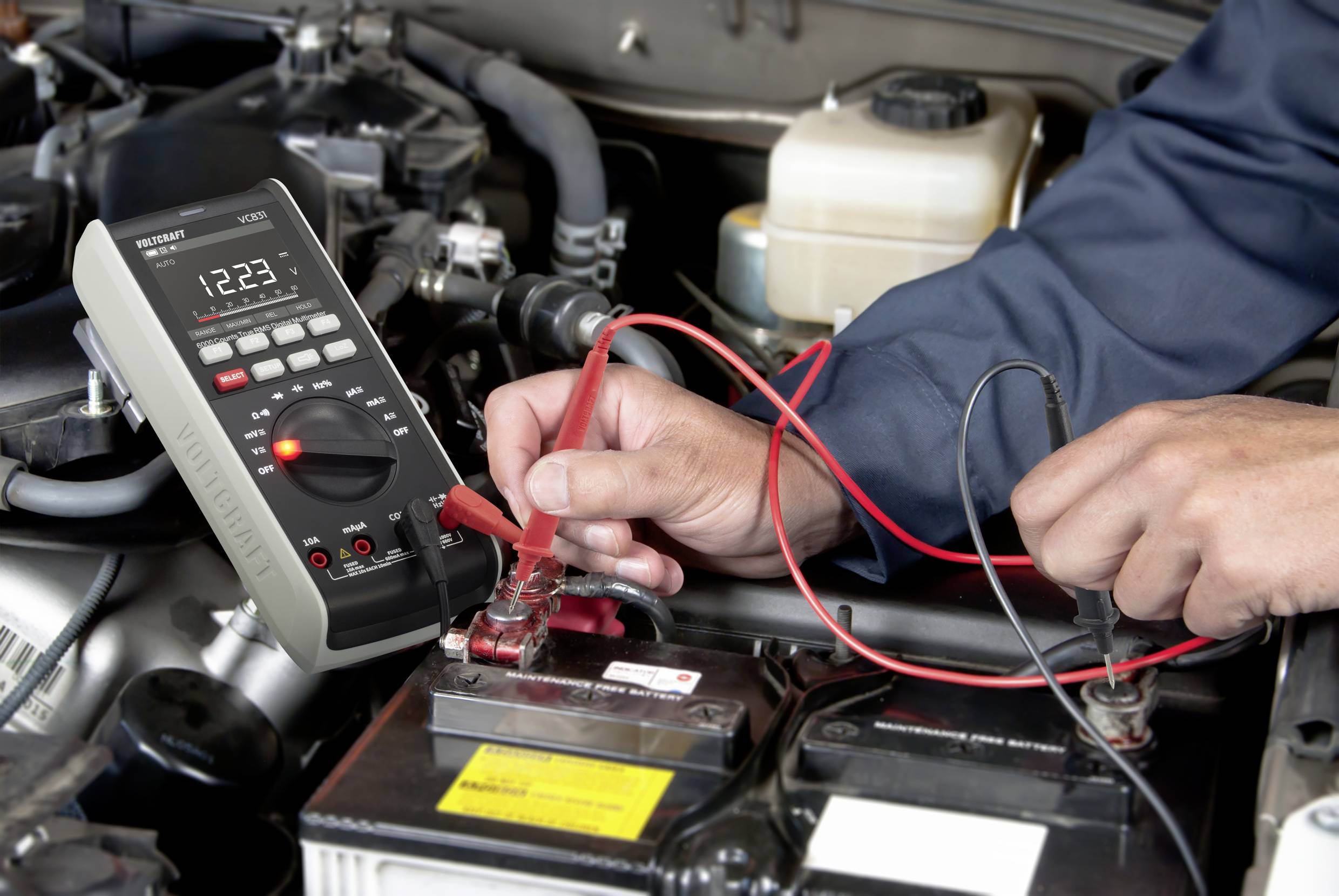 Osoba meria napätie autobatérie digitálnym multimetrom v motorovom priestore. Záber je zameraný na ruky a meracie zariadenie.