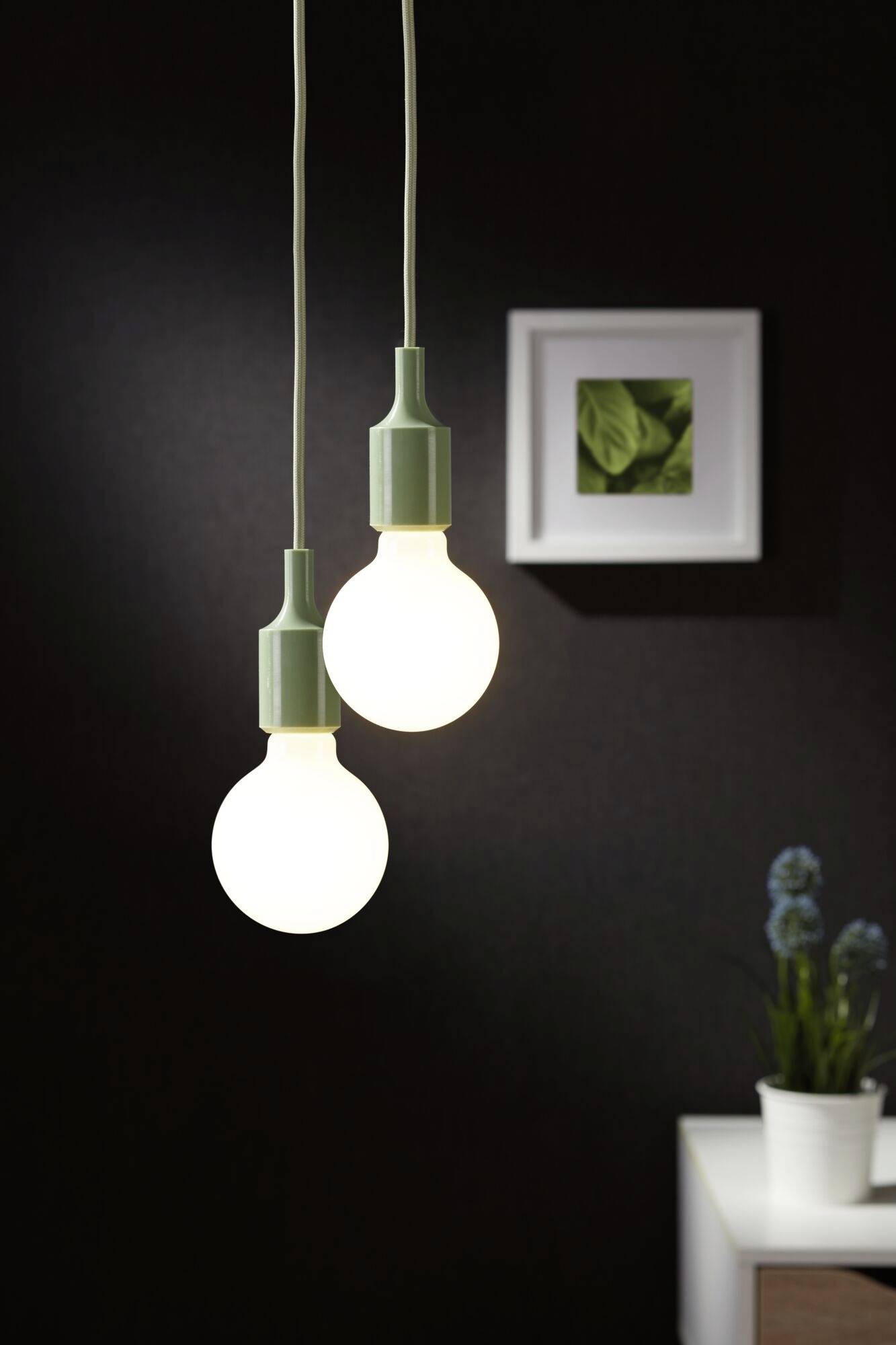 Dve visiacie žiarovky svietia v popredí tmavej miestnosti. V pozadí je zarámovaný obraz a rastlina na stole.