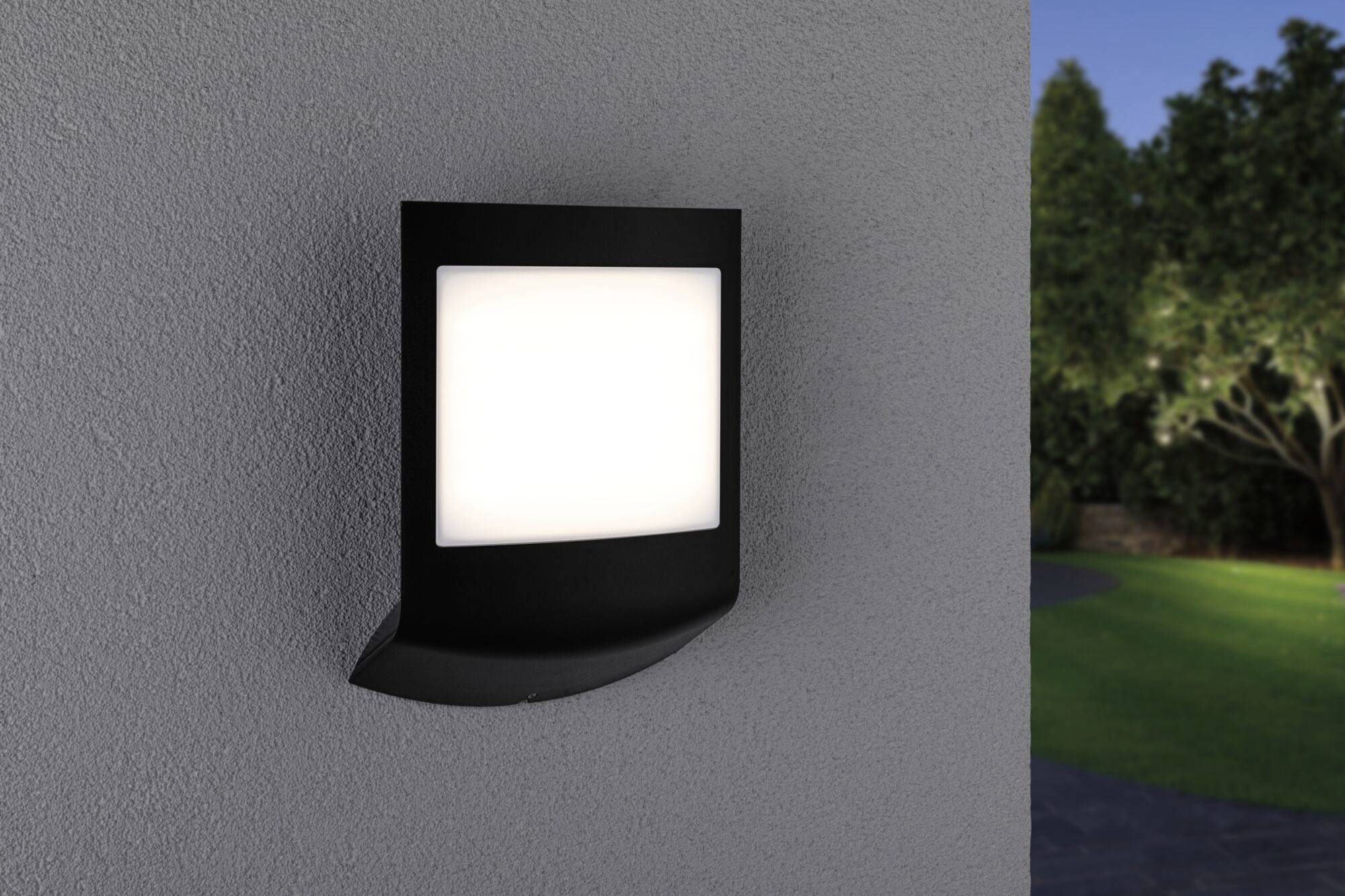 Nástenná lampa na sivej stene vyžaruje jasné svetlo a osvetľuje záhradu v pozadí s látím a trávnikom.