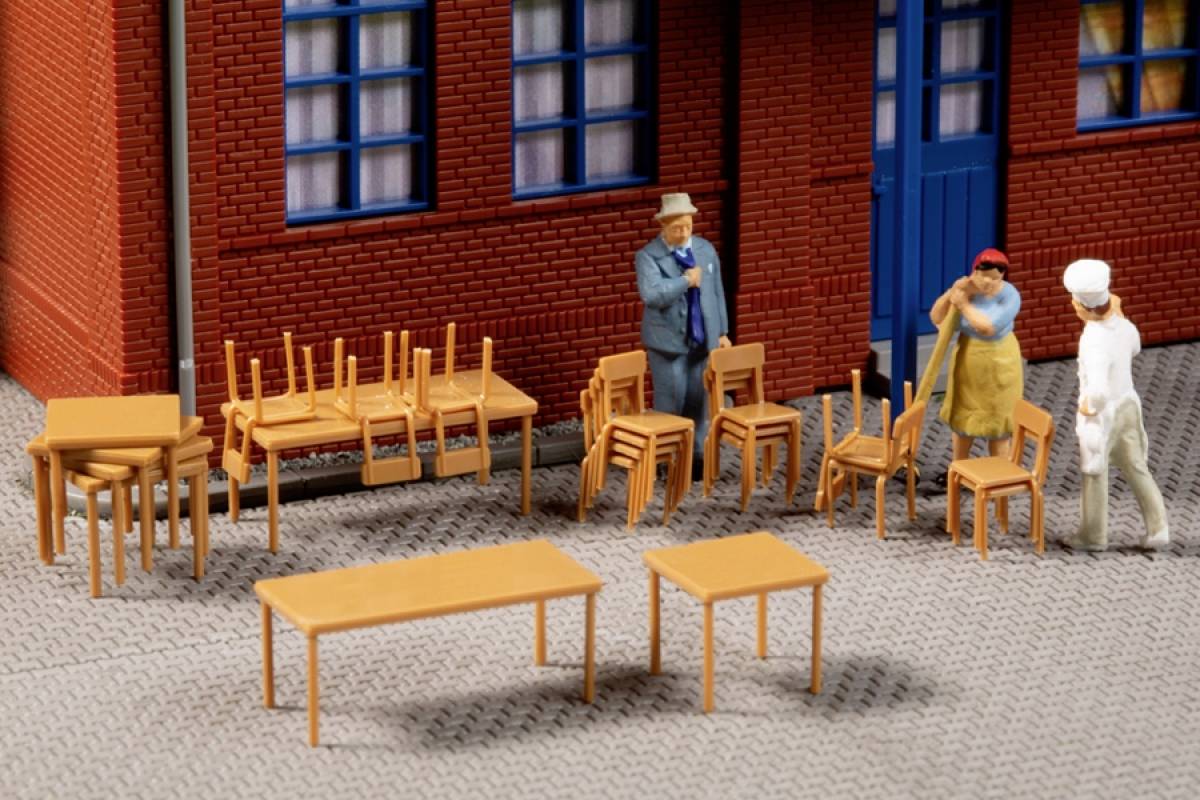 Miniaturový model zobrazuje tri figúry diskutujúce pred budovou. Vedľa nich sú poukladané stoličky a stôl.