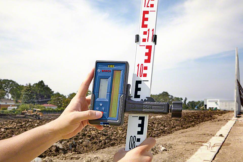 Osoba meria výškovú lať digitálnym nivelačným prístrojom na stavenisku. V pozadí sú stromy a zemné práce.