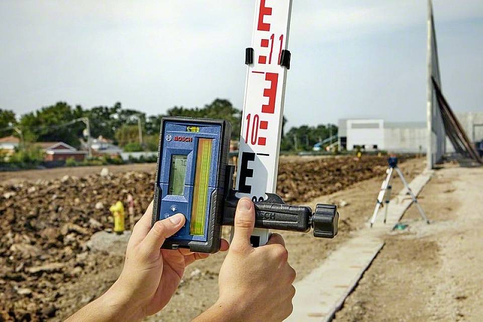 Osoba drží digitálny meradlo pri nivelačnej late na stavenisku za účelom určenia výšky terénu.