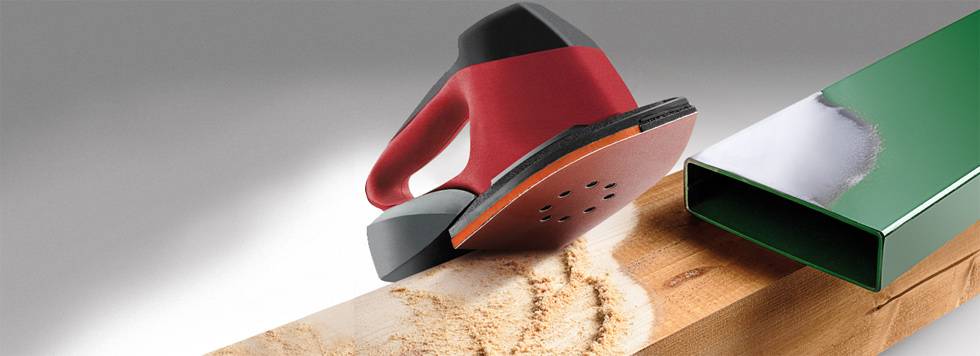 Červená excentrická brúska vyhladuje drevený povrch. Vedľa nej leží lakovaný kovový kus. V pozadí je sivý farebný prechod.