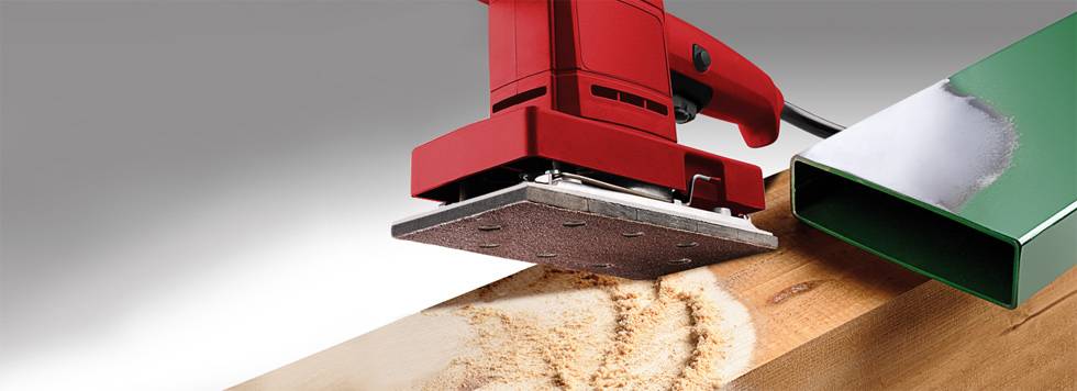 Červená vibračná brúska brúsi drevený povrch, pričom sa tvorí prach. Vedľa dreva leží zelená kovová súčiastka natretá farbou.