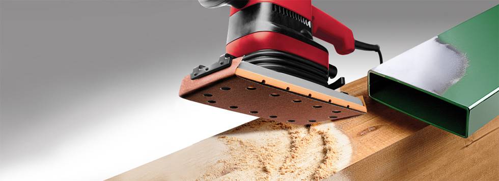 Červená elektrická brúska opracováva povrch dreveného trámu. Pozdĺž brúsenej plochy je viditeľný drevený prach.