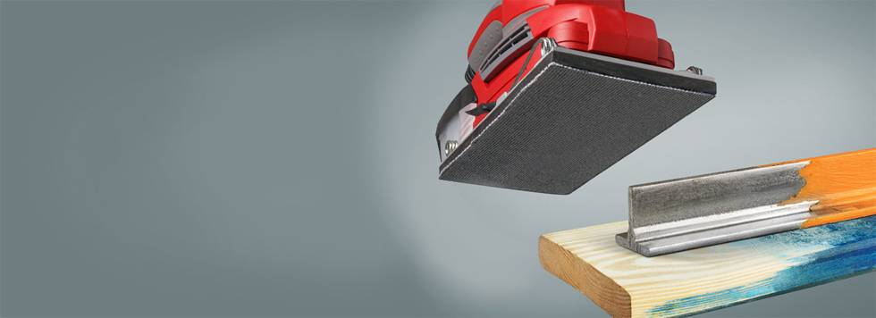 Červená brúska položená na drevenej doske a kovovej časti, pripravená na brúsenie. Pozadie je jednotvárne sivé.