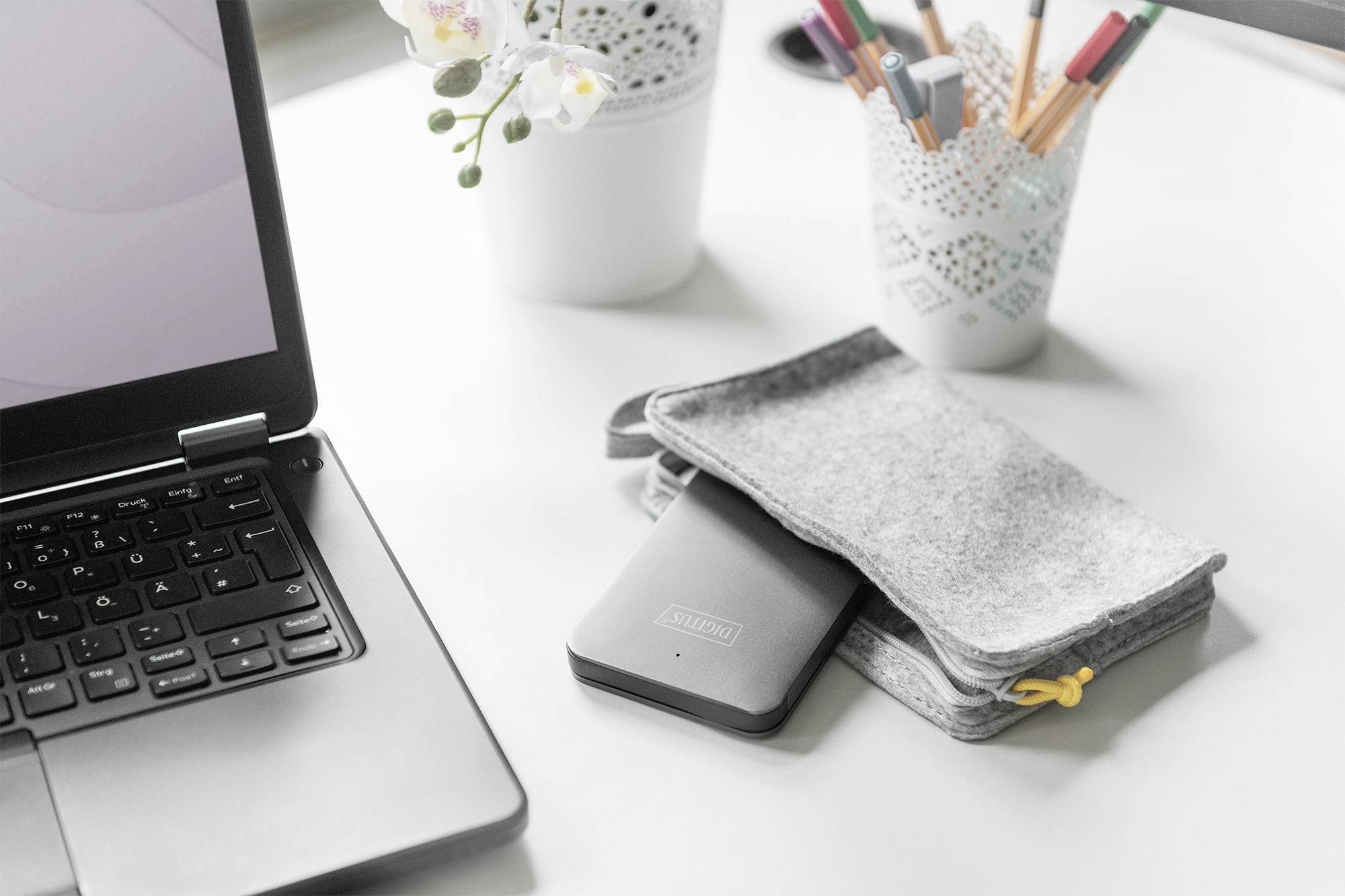 Otvorený laptop leží na bielom stole. Vedľa neho sú šedé plstené púzdro a externý pevný disk. V pozadí sú perá a rastlina.