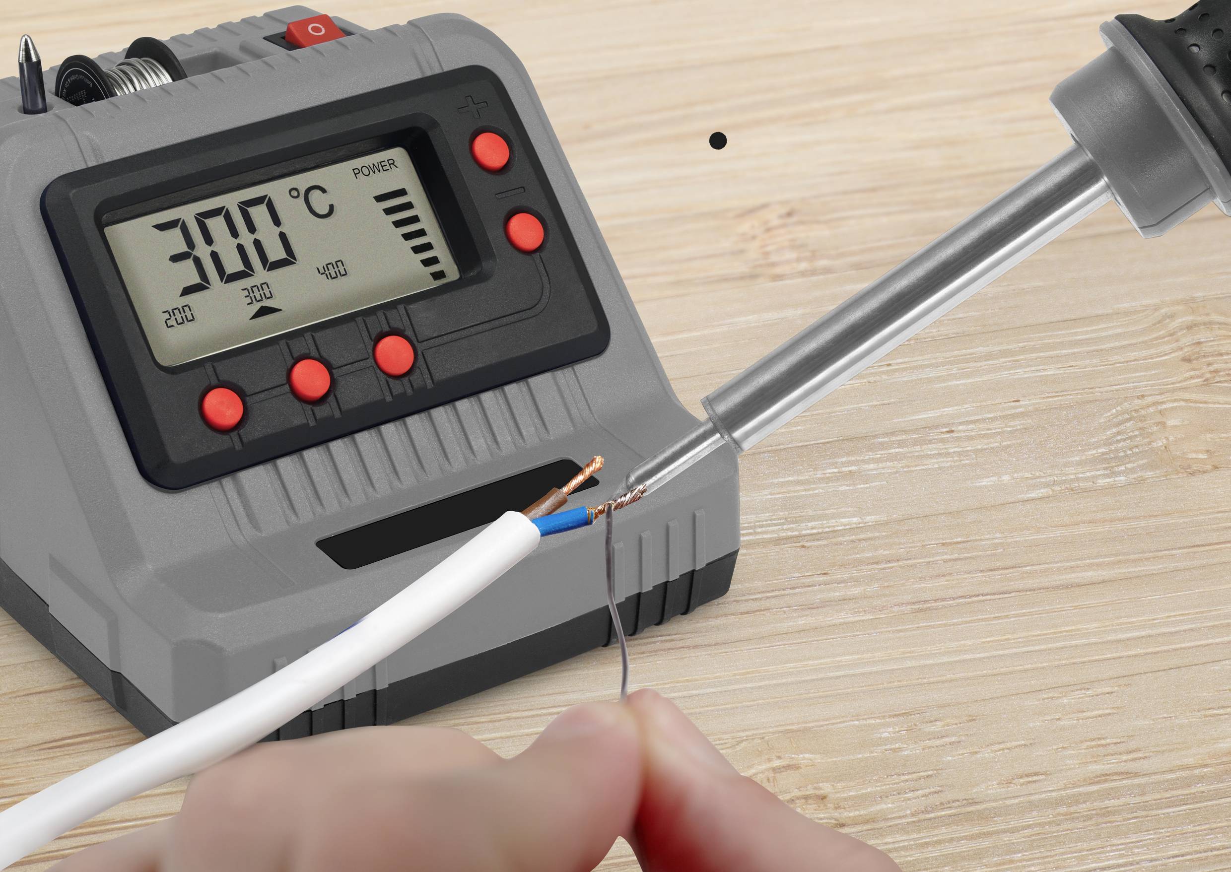 Spájkovačka s digitálnym displejom ukazuje 300°C. Niekto spájkuje konce káblov na svetlom drevenom stole.