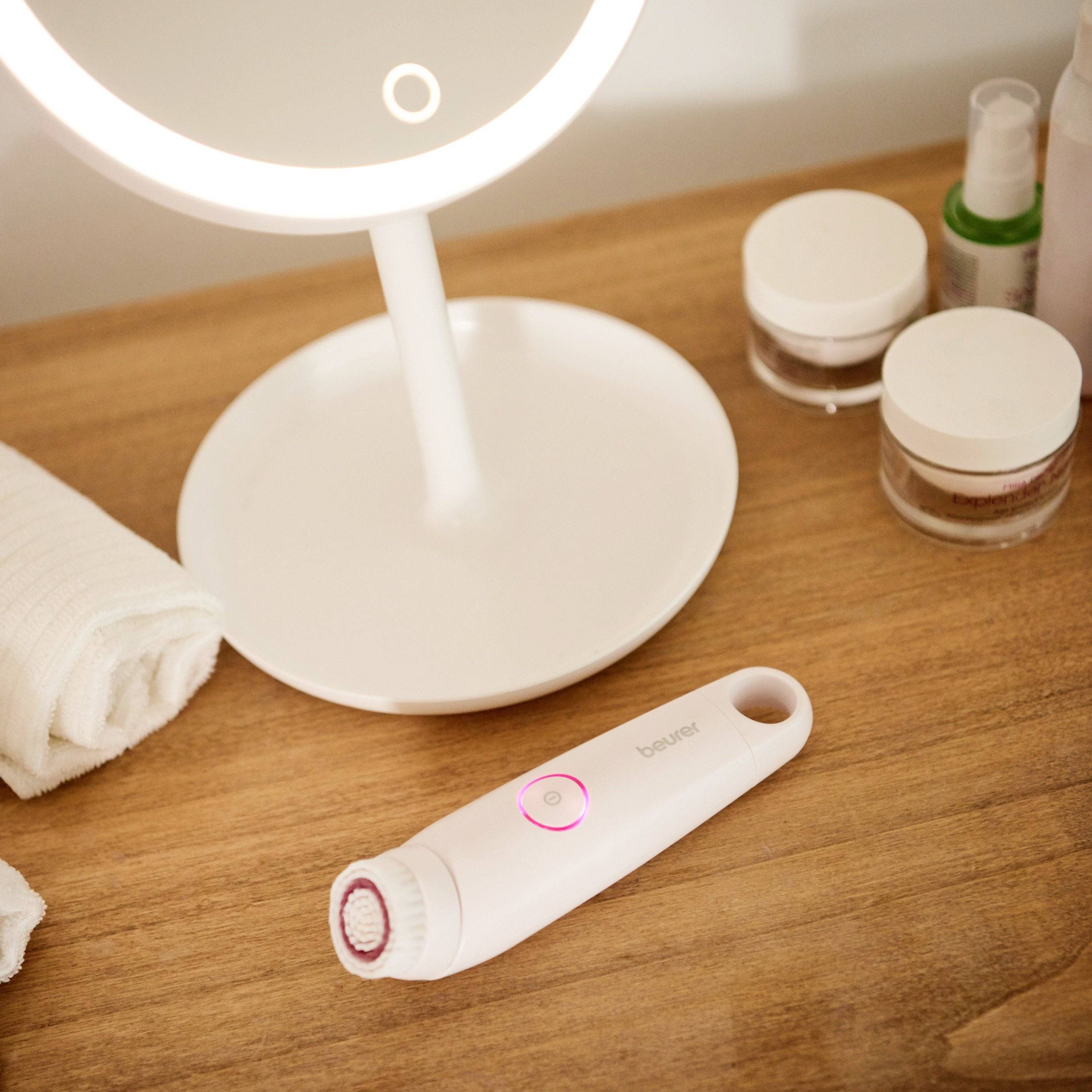 Osvetlené make-up zrkadlo na drevenom stole, vedľa leží elektrická čistiaca kefa. Na stole sú nádobky a uterák.