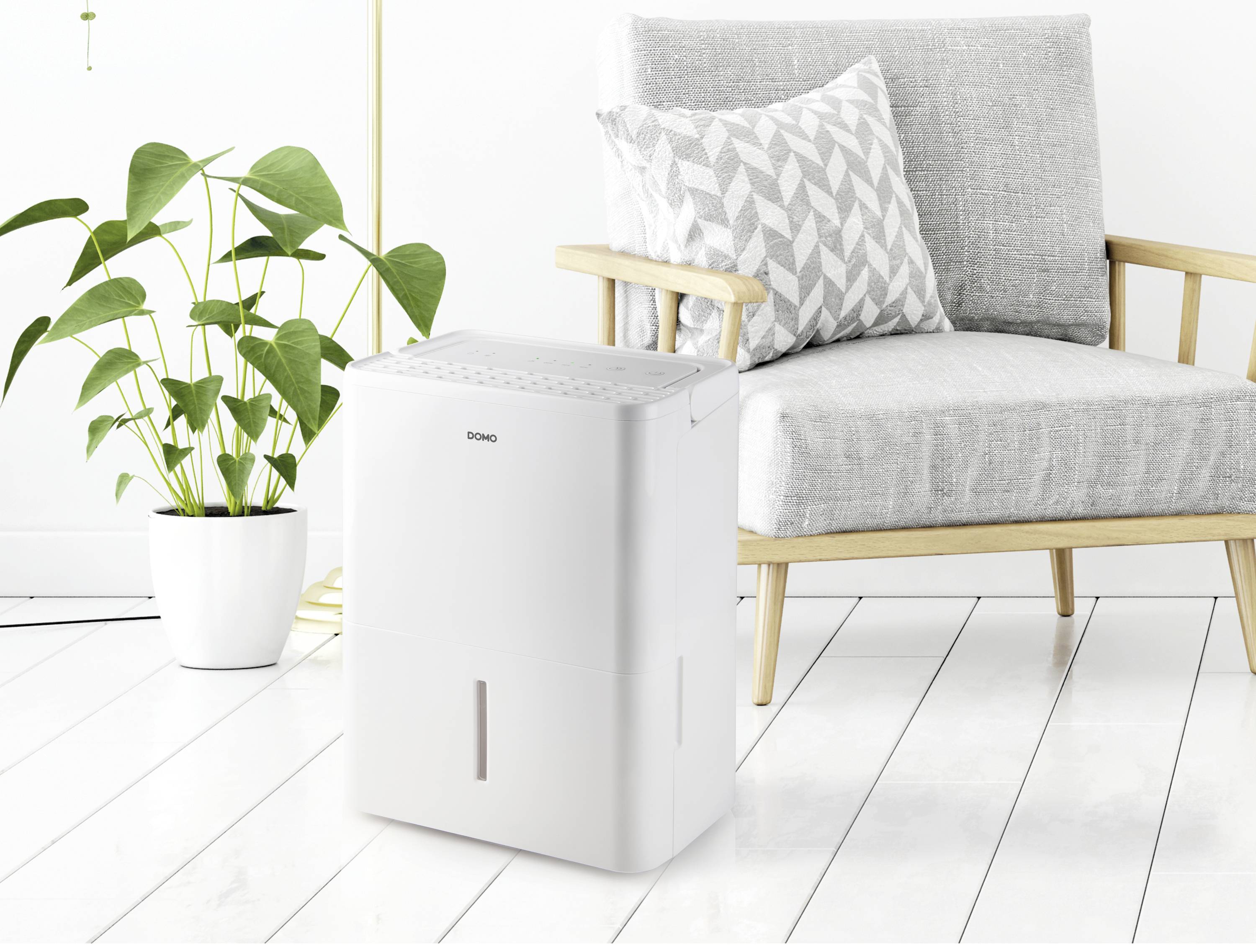 Biely odvlhčovač vzduchu stojí na svetlej drevenej podlahe vedľa sivého kresla a izbovej rastliny v modernej miestnosti.