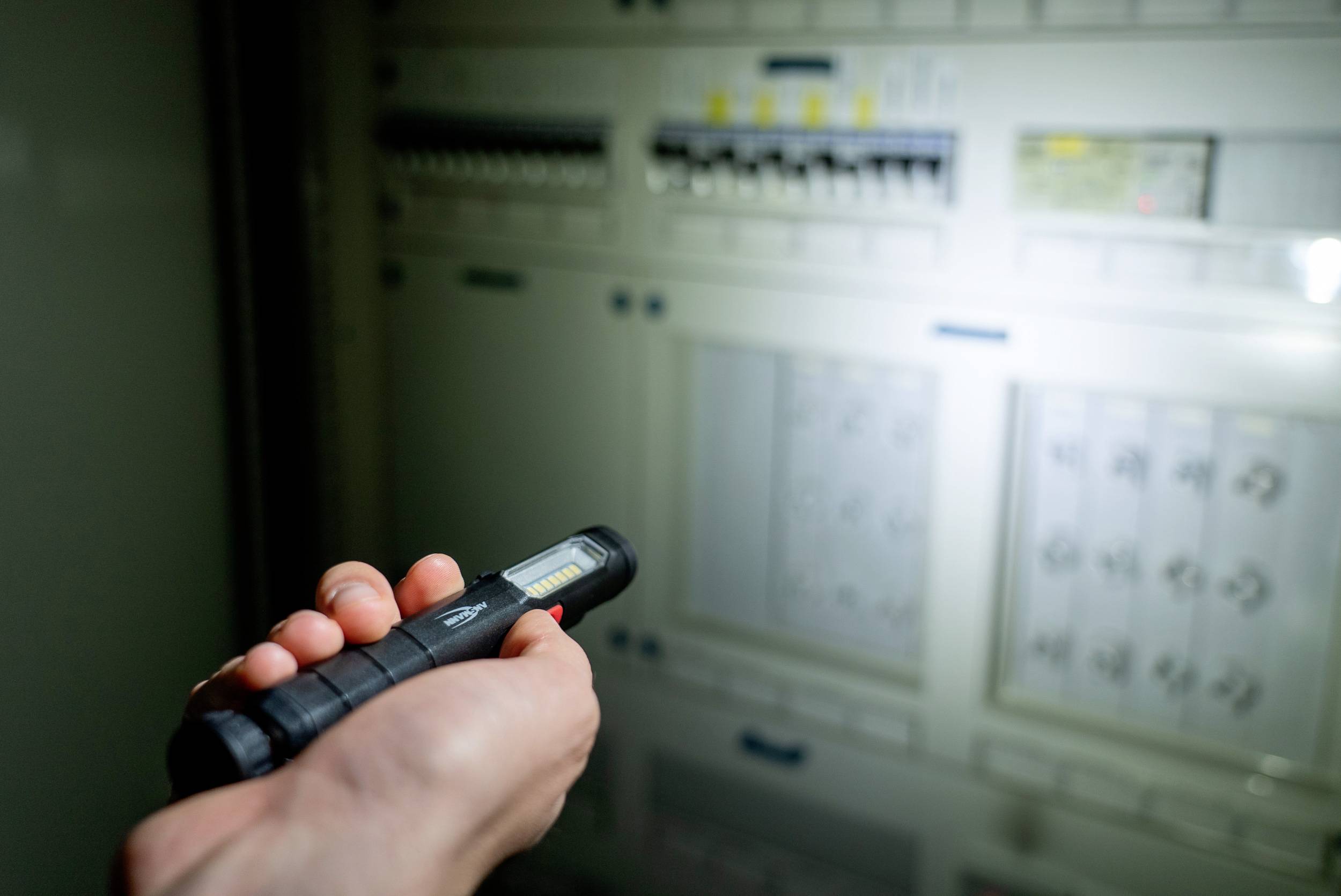 Ruka drží baterku a osvetľuje elektrický rozvádzač, na ktorom sú viditeľné viaceré spínače a indikátory.