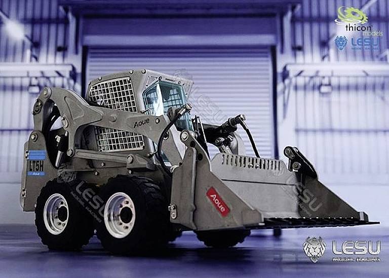 Strieborný, detailne spracovaný model bagra stojí v hale. Má šesť kolies a lyžicu vpredu. Na stroji sú umiestnené logá.