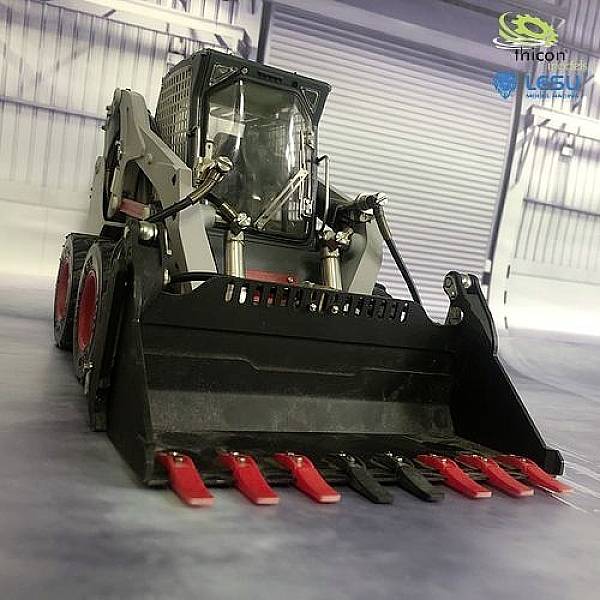 Kompaktný nakladač v priemyselnej hale, zobrazený spredu so zdvíhacou lyžicou a hydraulickými ramenami. V pozadí je rolovacia brána.
