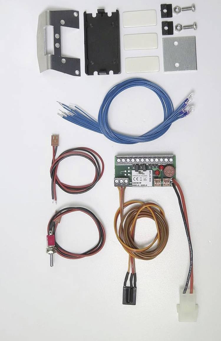 Elektronický stavebnicový set na bielom pozadí. Obsahuje káble, spínače, dosku plošných spojov a upevňovacie prvky. Vhodný pre DIY projekty.