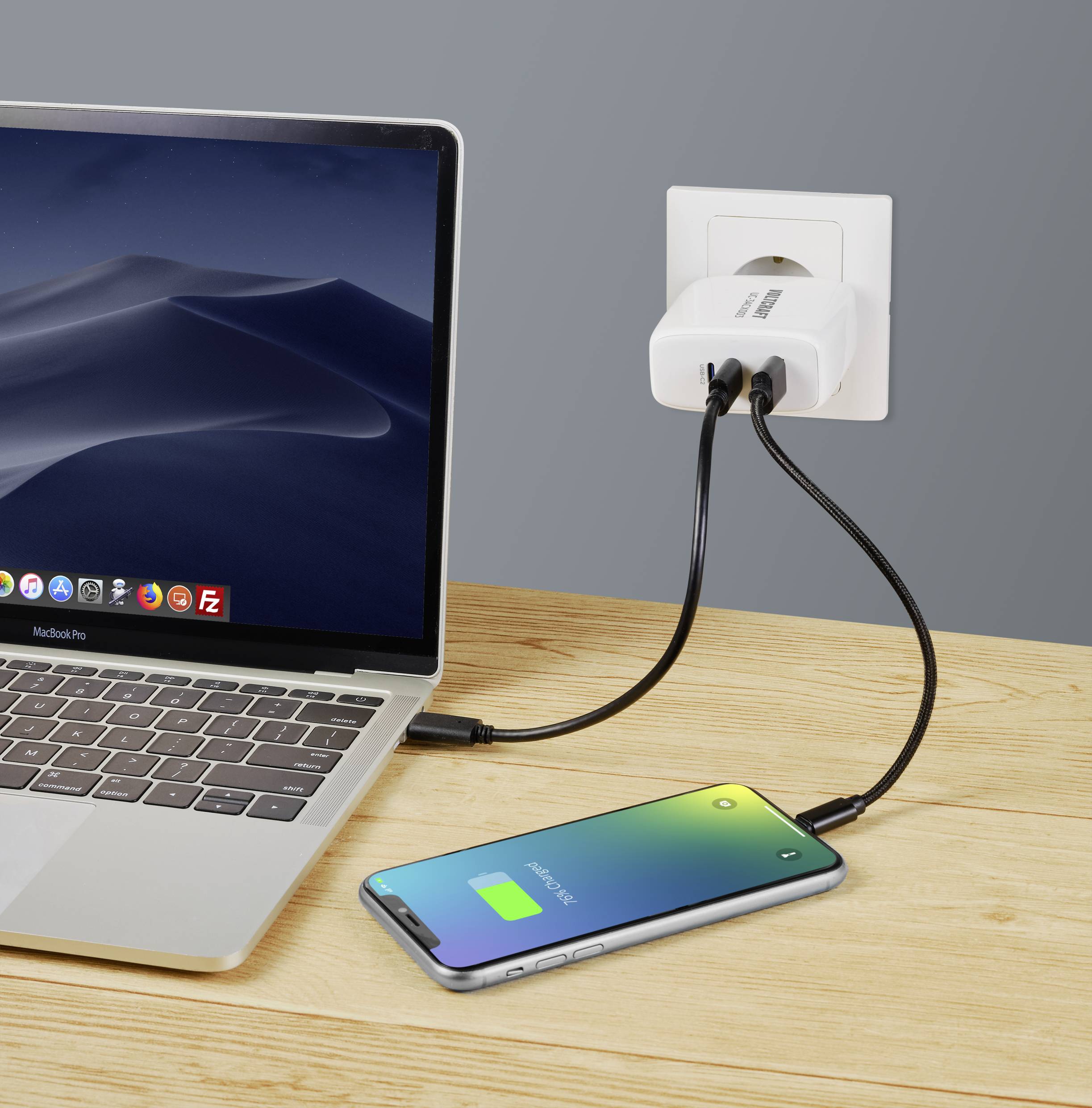 Laptop a smartfón na drevenom stole. Obe zariadenia sú pripojené k nabíjačke v elektrickej zásuvke. Smartfón zobrazuje zelený stav batérie.