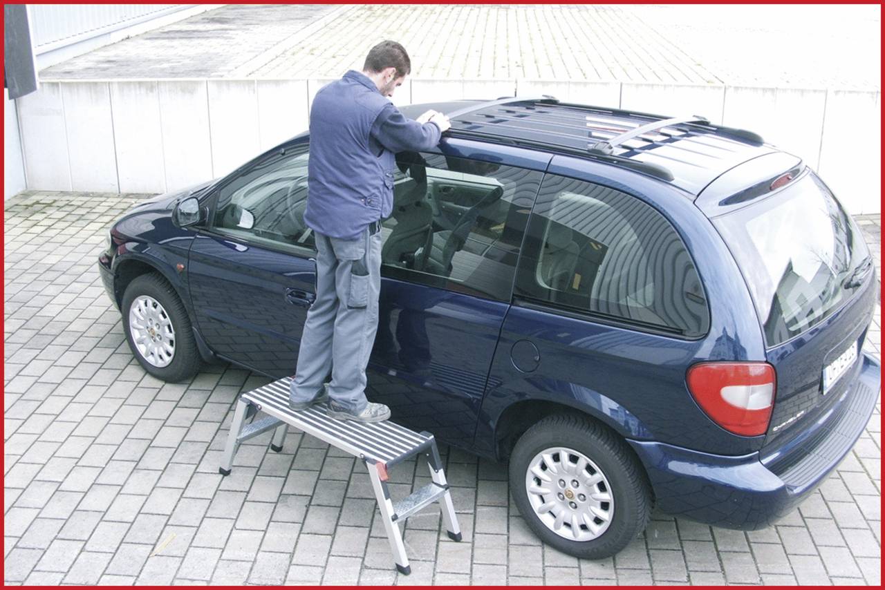 Muž stojí na rebríku vedľa modrého auta a pracuje na streche, na dláždenej ploche pred budovou.