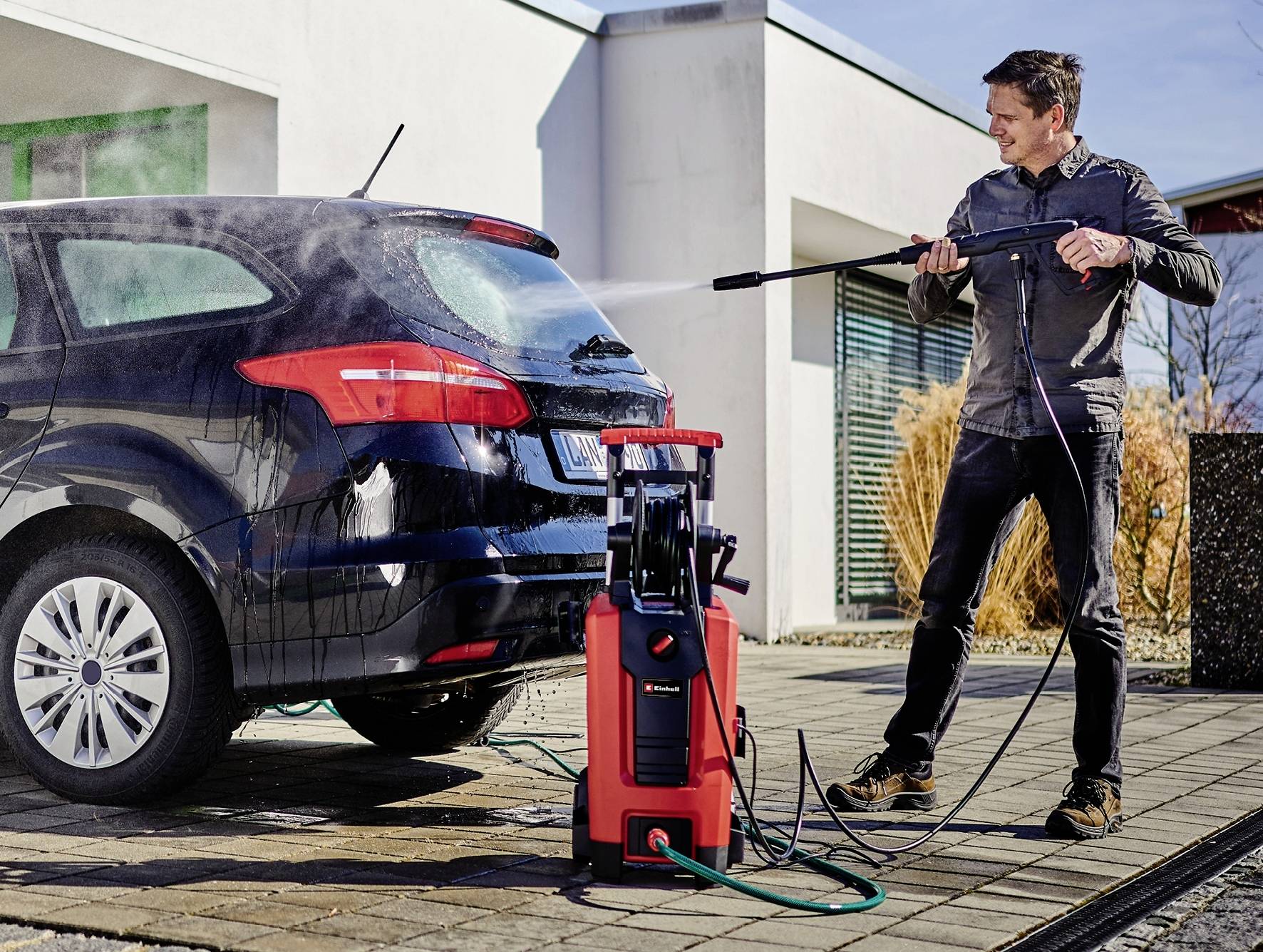 Muž čistí auto vysokotlakovým čističom na príjazdovej ceste pred modernou budovou.