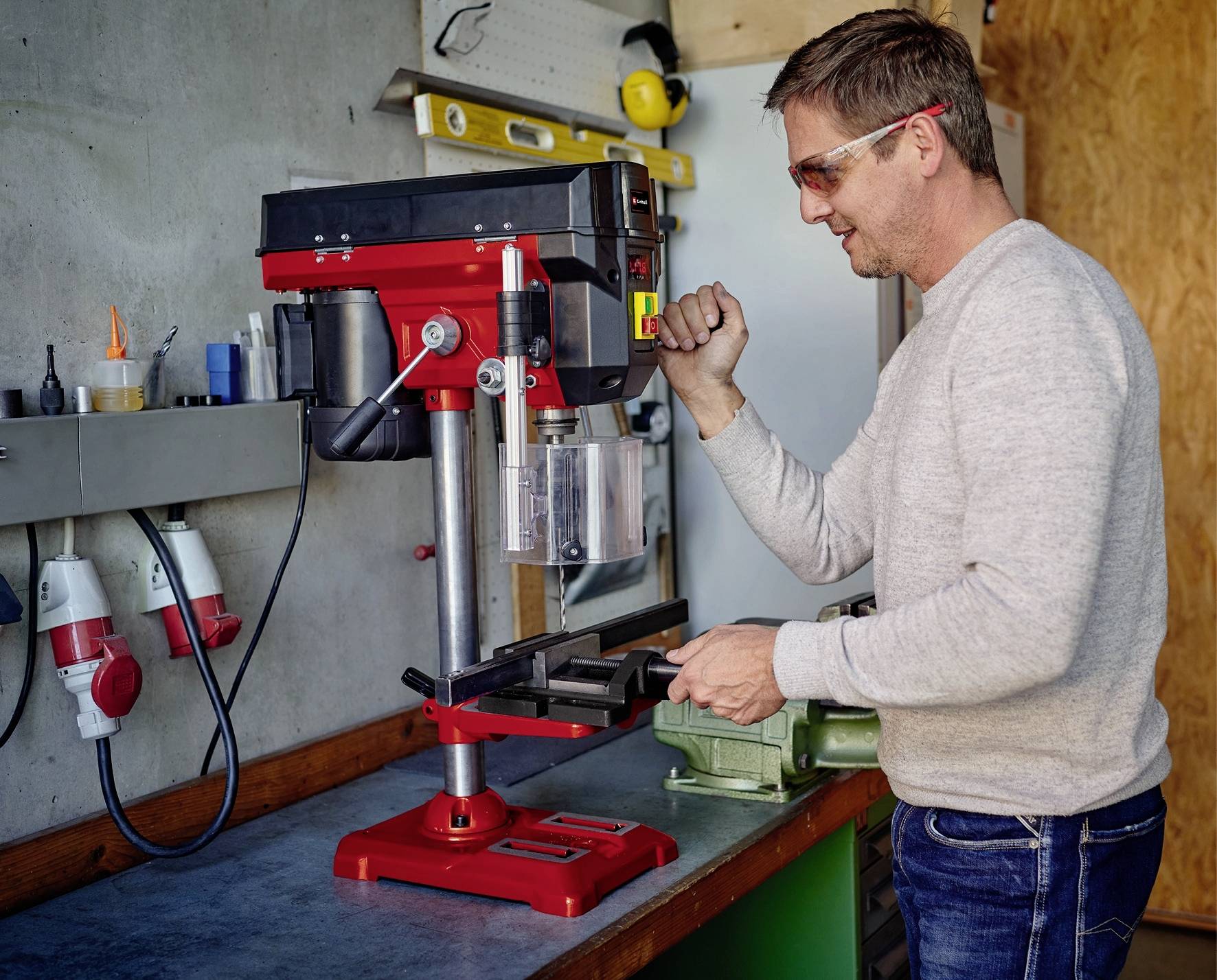 Muž obsluhuje stolovú vŕtačku v dielni. Má na sebe ochranné okuliare a pravou rukou drží páku.