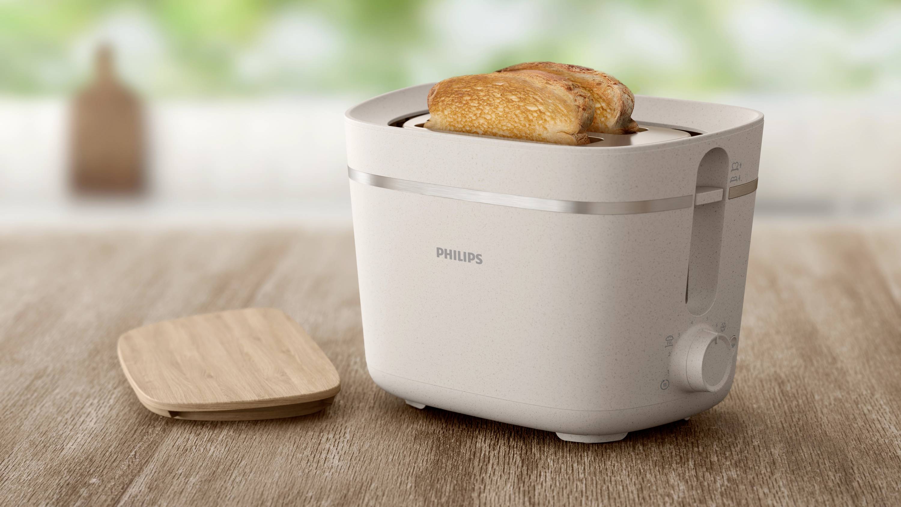 Toaster značky Philips s dvoma opečenými toastnými plátkami na drevenej doske. V rozmazanom pozadí ďalší kuchynský doplnok.
