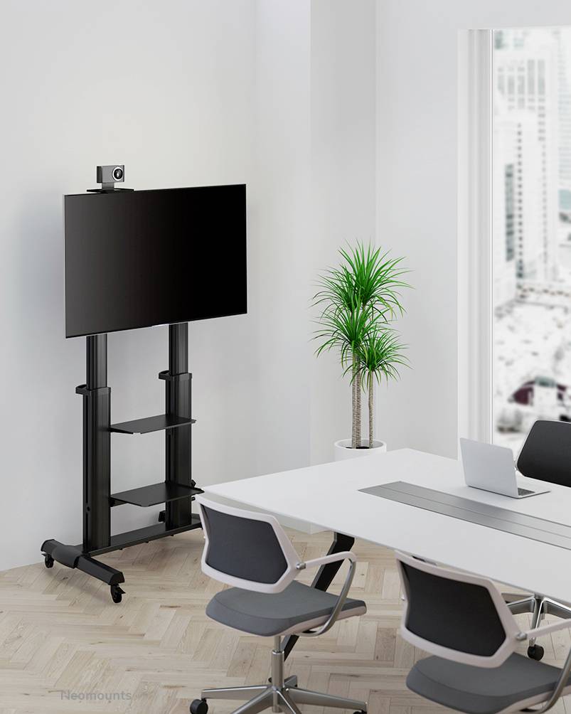 Moderná konferenčná miestnosť s bielym stolom, tromi kancelárskymi stoličkami a veľkou plochou obrazovkou na mobilnom stojane. V pozadí stojí zelená rastlina.