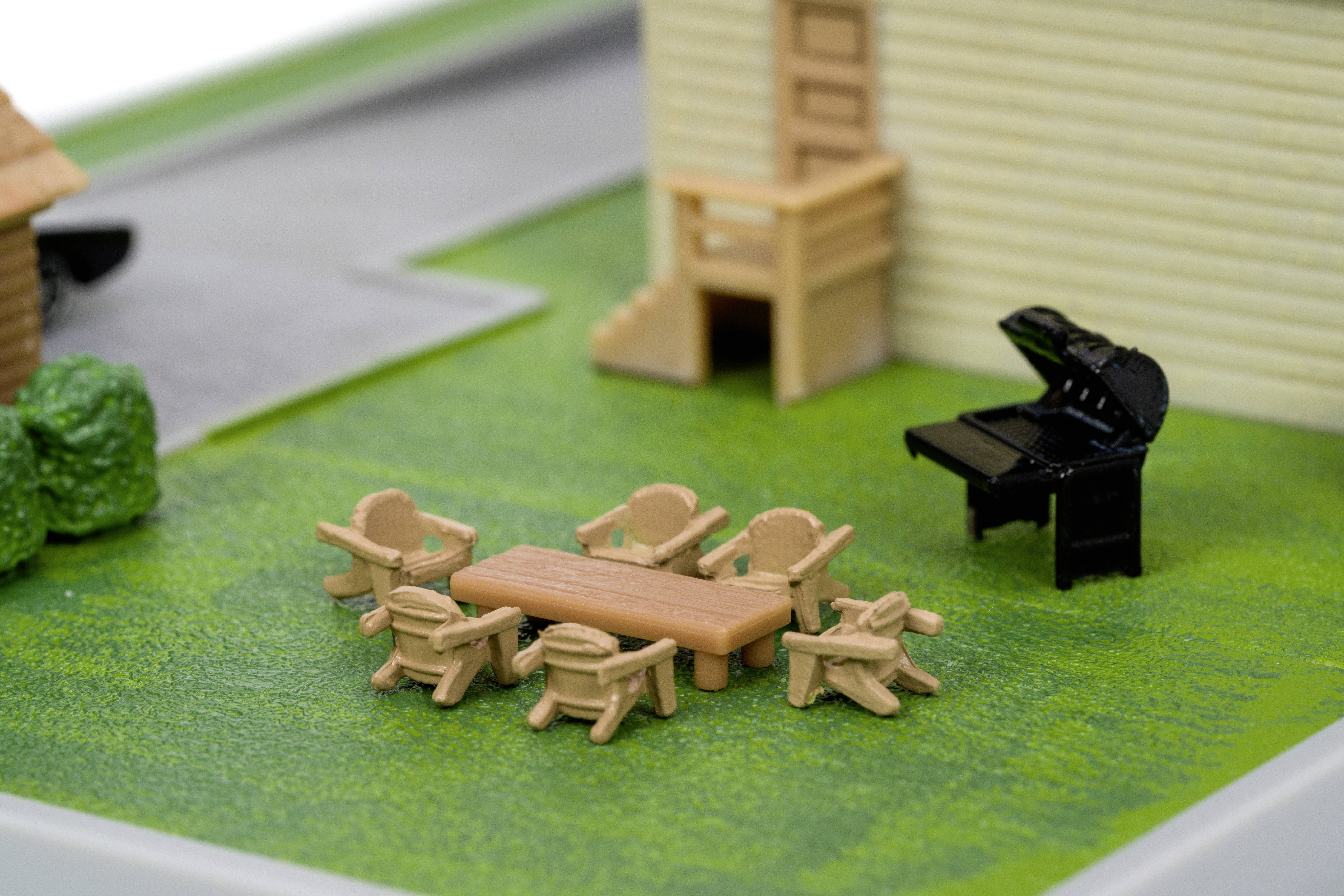 Miniatúrna terénna scéna so stolom, stoličkami a klavírom na zelenej tráve vedľa budovy.