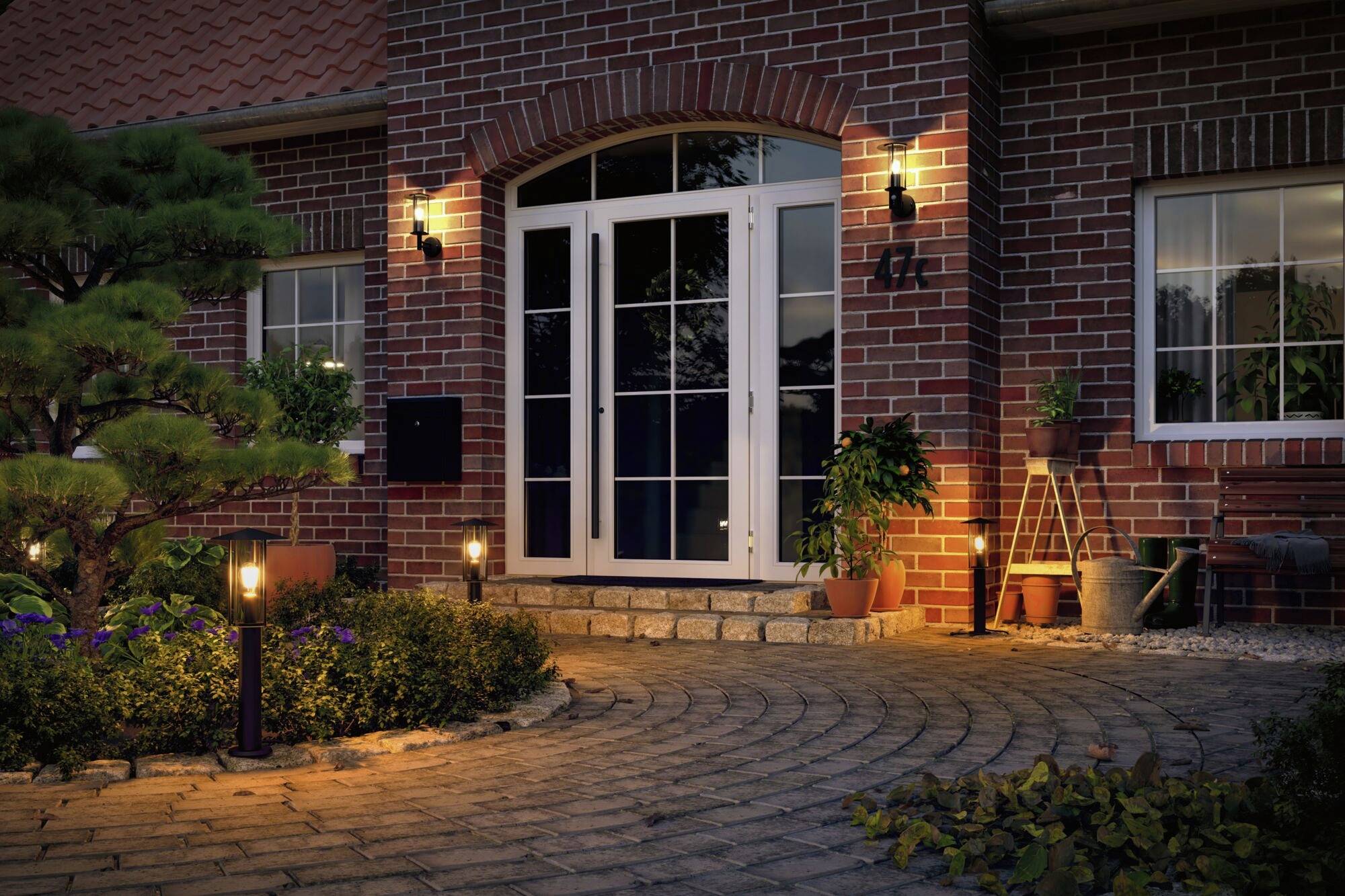 Osvetlený vstup do domu s veľkými bielymi dverami, obklopený kamennou dlažbou a kvetináčmi, za súmraku.