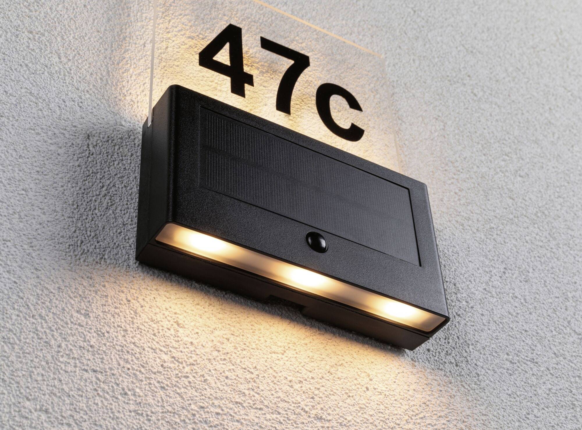 Na sivej stene visí čierna vonkajšia lampa s tromi svietiacimi žiarovkami pod tabuľou s nápisom '47C'.