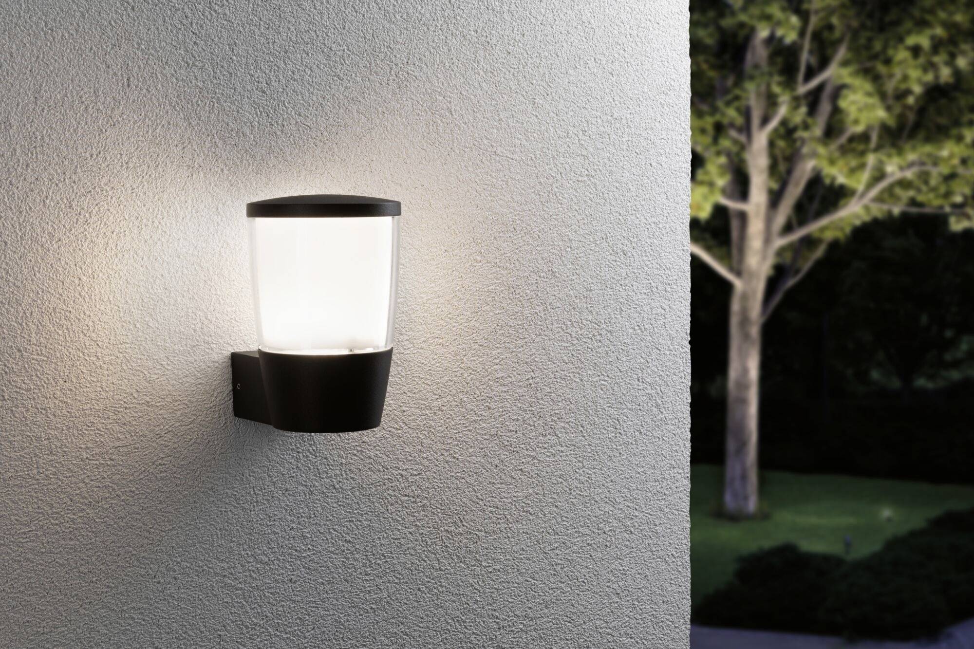 Vonkajšia lampa svieti na stene a osvetľuje záhradu porastenú stromami v pozadí.