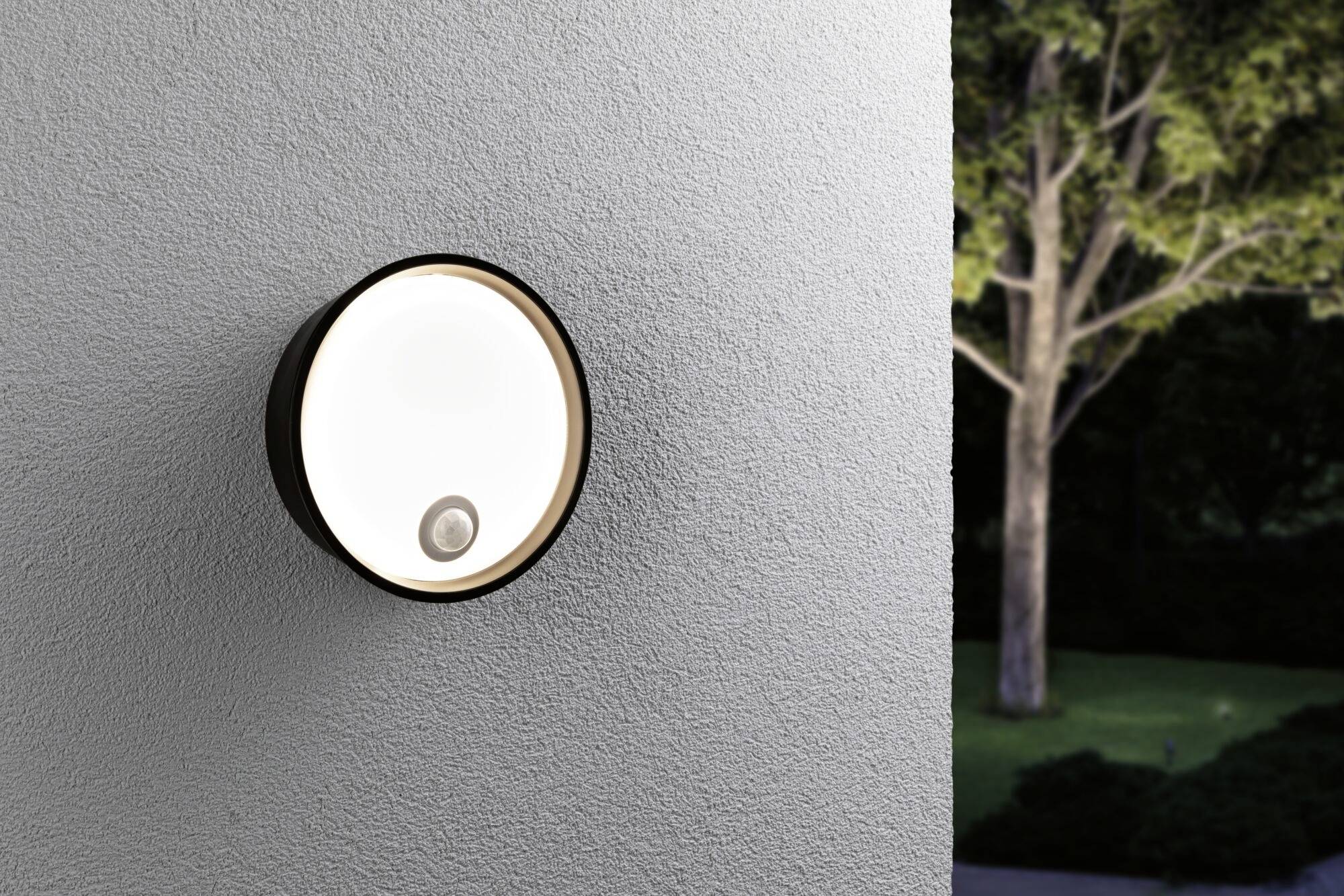 Nástená lampa s pohybovým senzorom na sivej stene, osvetlená záhrada v pozadí. Osvetlenie pre bezpečnosť v exteriéri.