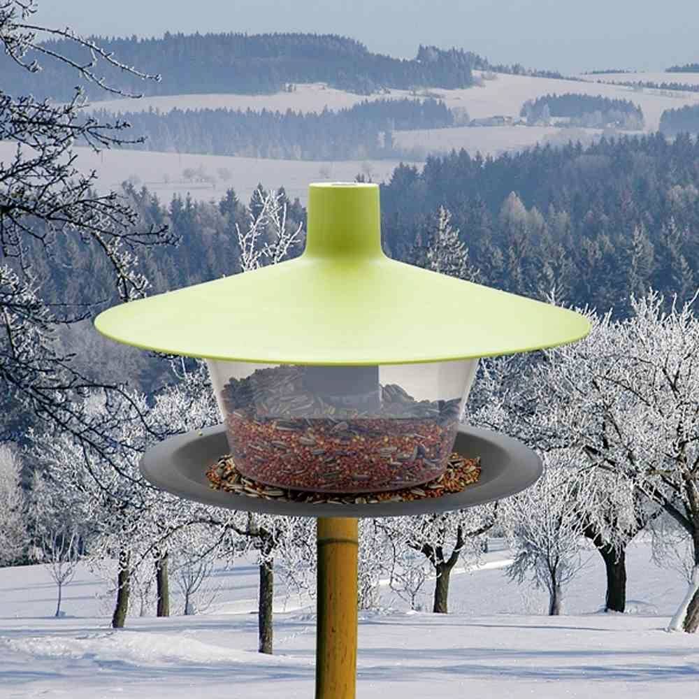 Zelený vtáčí domček s krmivom, v zasneženej lesnej krajine v pozadí. Žiadne viditeľné vtáky. Zimná snehová krajina.