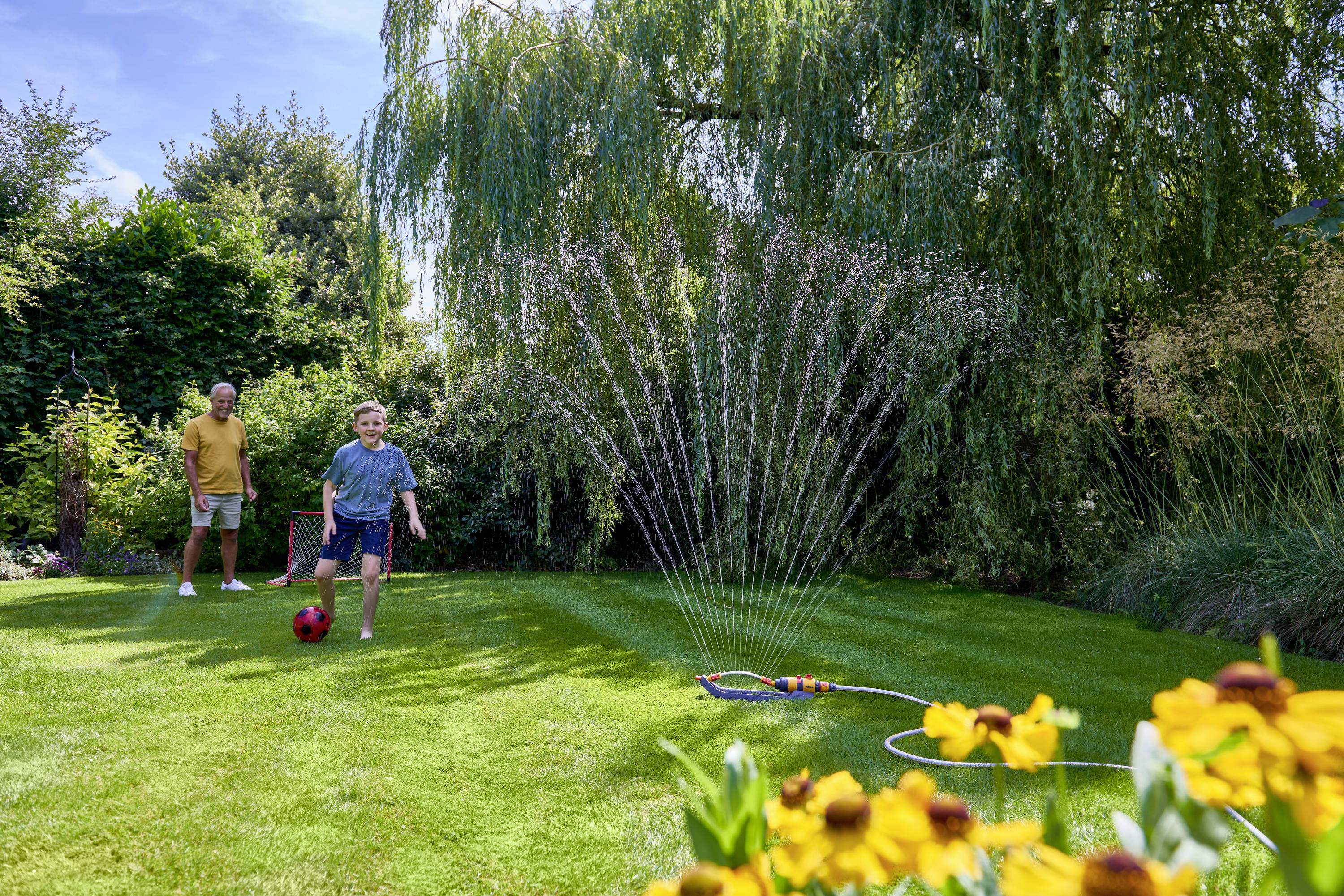 Chlapec sa hrá s červenou loptou v záhrade, pričom ho starší muž pozoruje. Rozprašovač zavlažuje trávnik. Kvety v popredí.