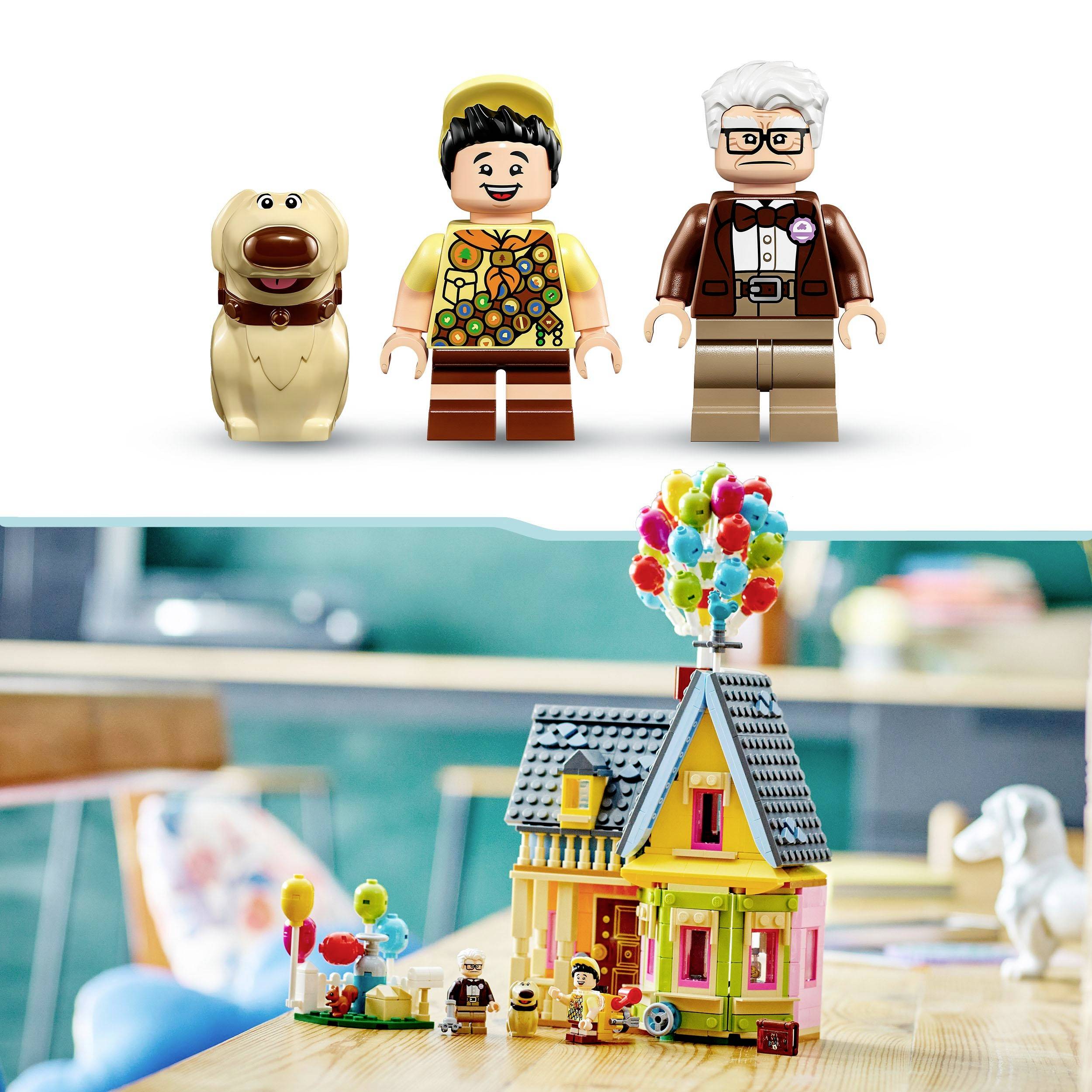 Tri figúrky Lego stoja pred miniatúrnym modelom farebného domu s balónmi. V pozadí sú rozmazané kontúry nábytku.