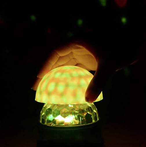 Ruka sa dotýka svietiacej lampy v tvare huby, ktorá vytvára zelené a červené svetelné efekty na tmavom povrchu.