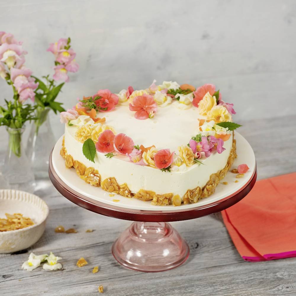 Na ružovom podnose stojí okrúhla torta ozdobená farebnými jedlými kvetmi, vedľa vázy s kvetmi.