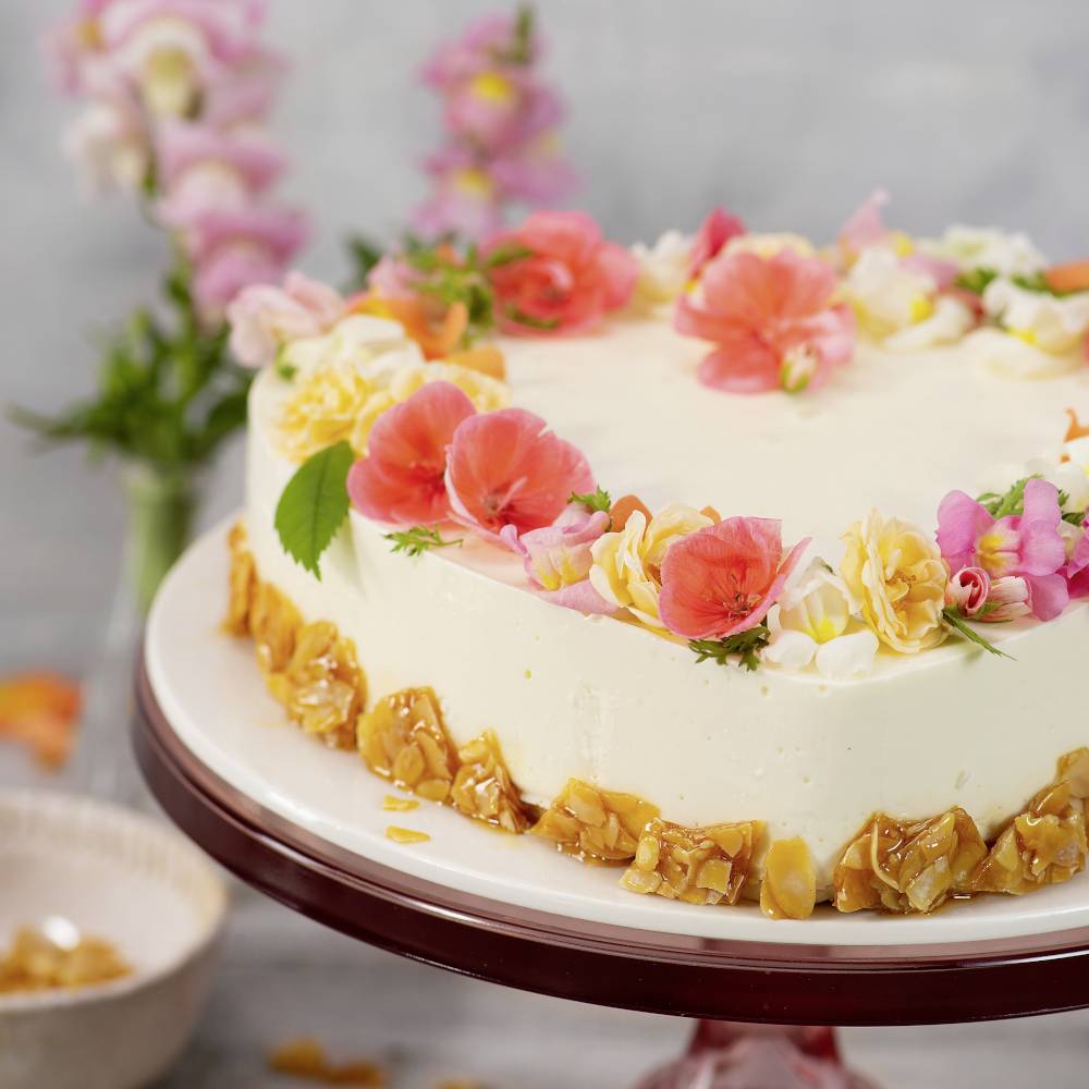 Okrúhla torta so svetlým krémom, ozdobená farebnými kvetmi a kúskami karamelu na lesklom tanieri. V pozadí kvety.