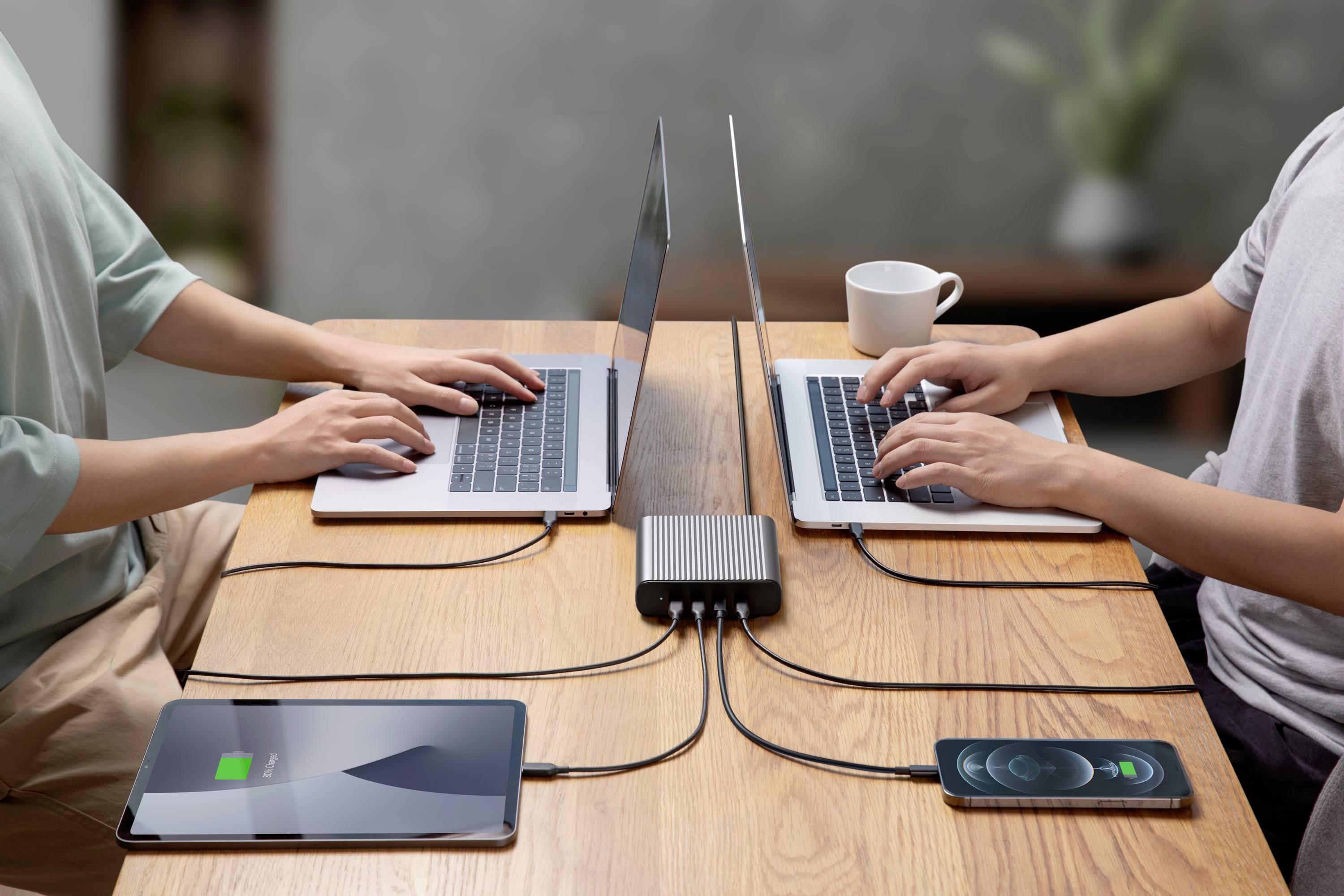 Dve osoby pracujú na laptopoch pri stole, pripojené k nabíjačke. Tablet a smartfón sa nabíjajú súčasne.