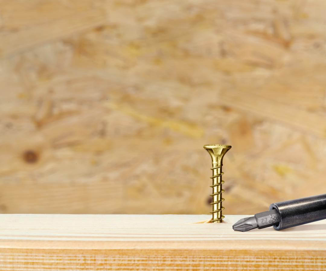 Skrutkovač sa opiera o vyčnievajúci zlatý skrutku na svetlom dreve, s rozmazaným dreveným pozadím dosiek.