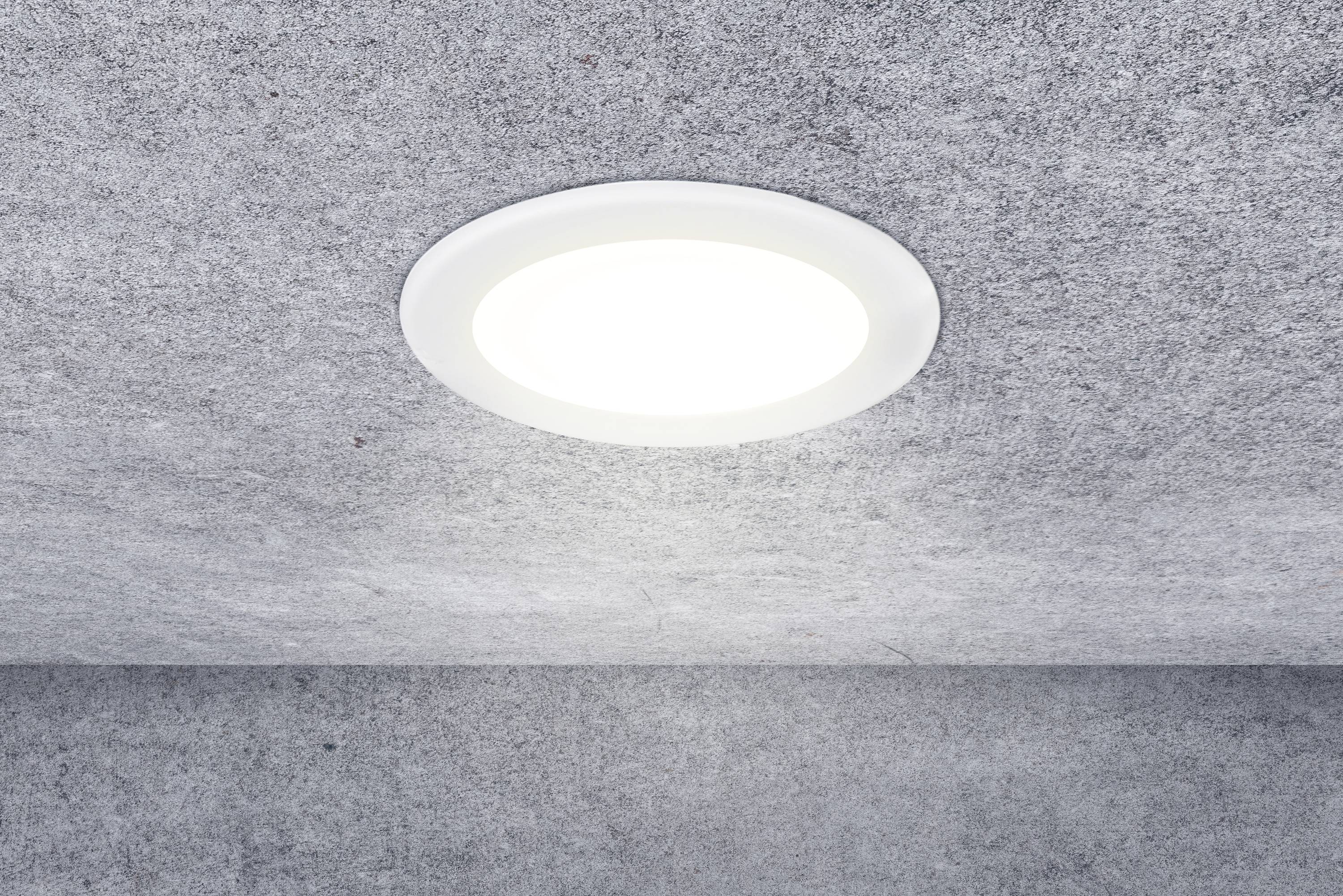 Biela, okrúhla stropná lampa svieti v miestnosti s šedou štruktúrou stropu a stien.