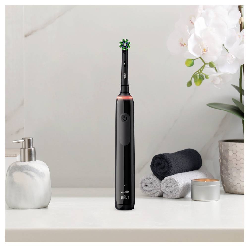 Čierna elektrická zubná kefka stojí kolmo na kúpeľňovom pulte, v pozadí sú zvinené uteráky, dávkovač mydla a biele orchidey.